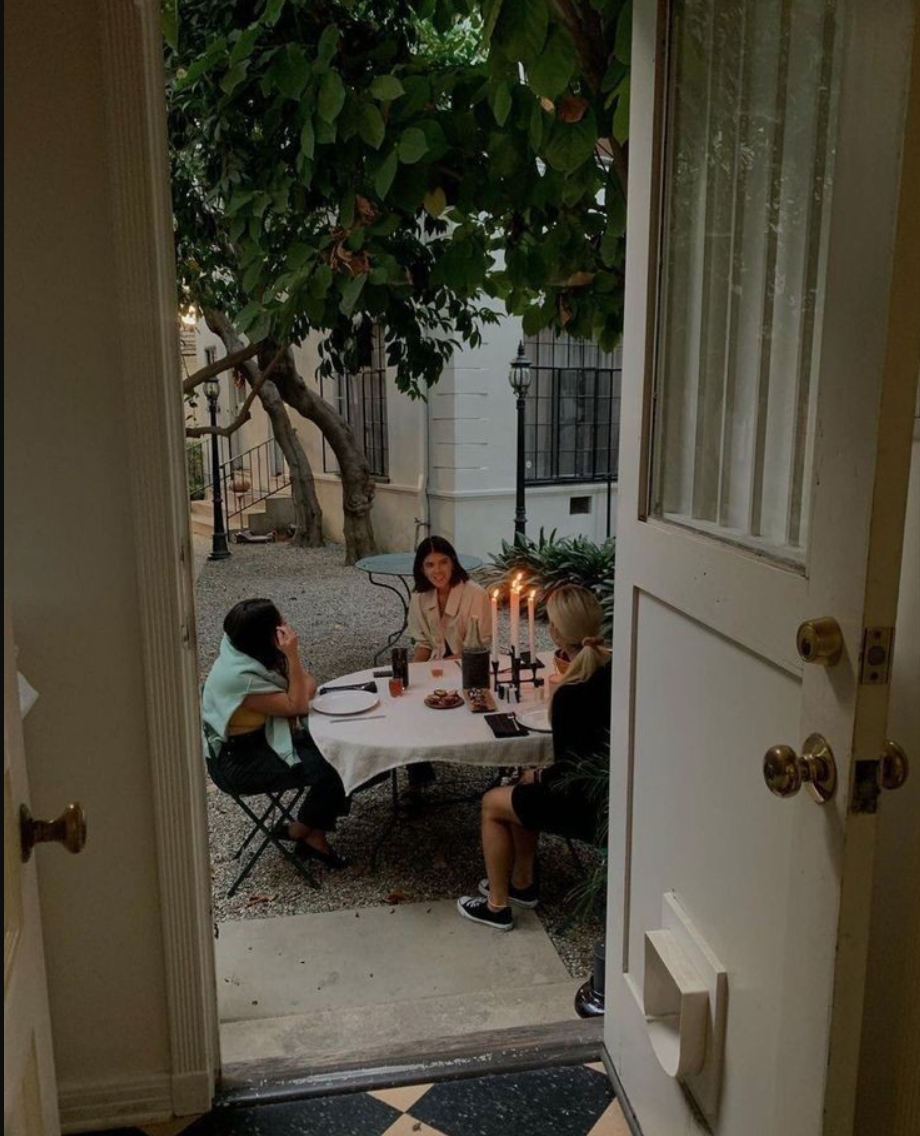 Black and white photo of three women with long dark hair, covering their eyes with their hands, standing in front of a wooden fence surrounded by flowers and foliage.