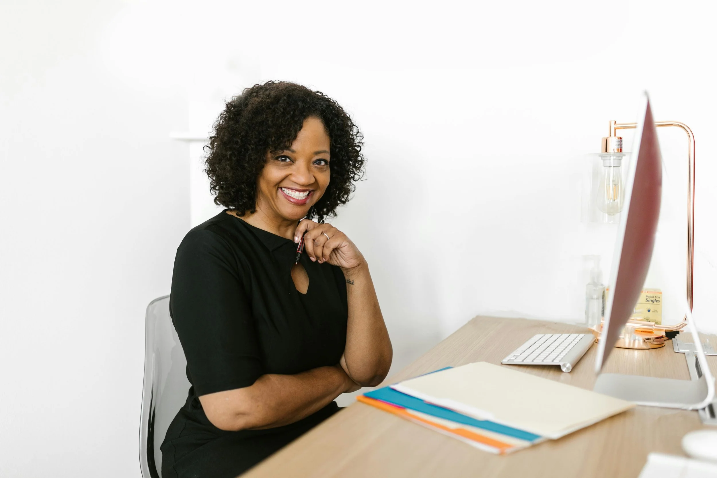 A smiling woman with curly dark hair sitting at a desk with a computer, colorful folders, and a pen in hand, in a bright, minimalist office.