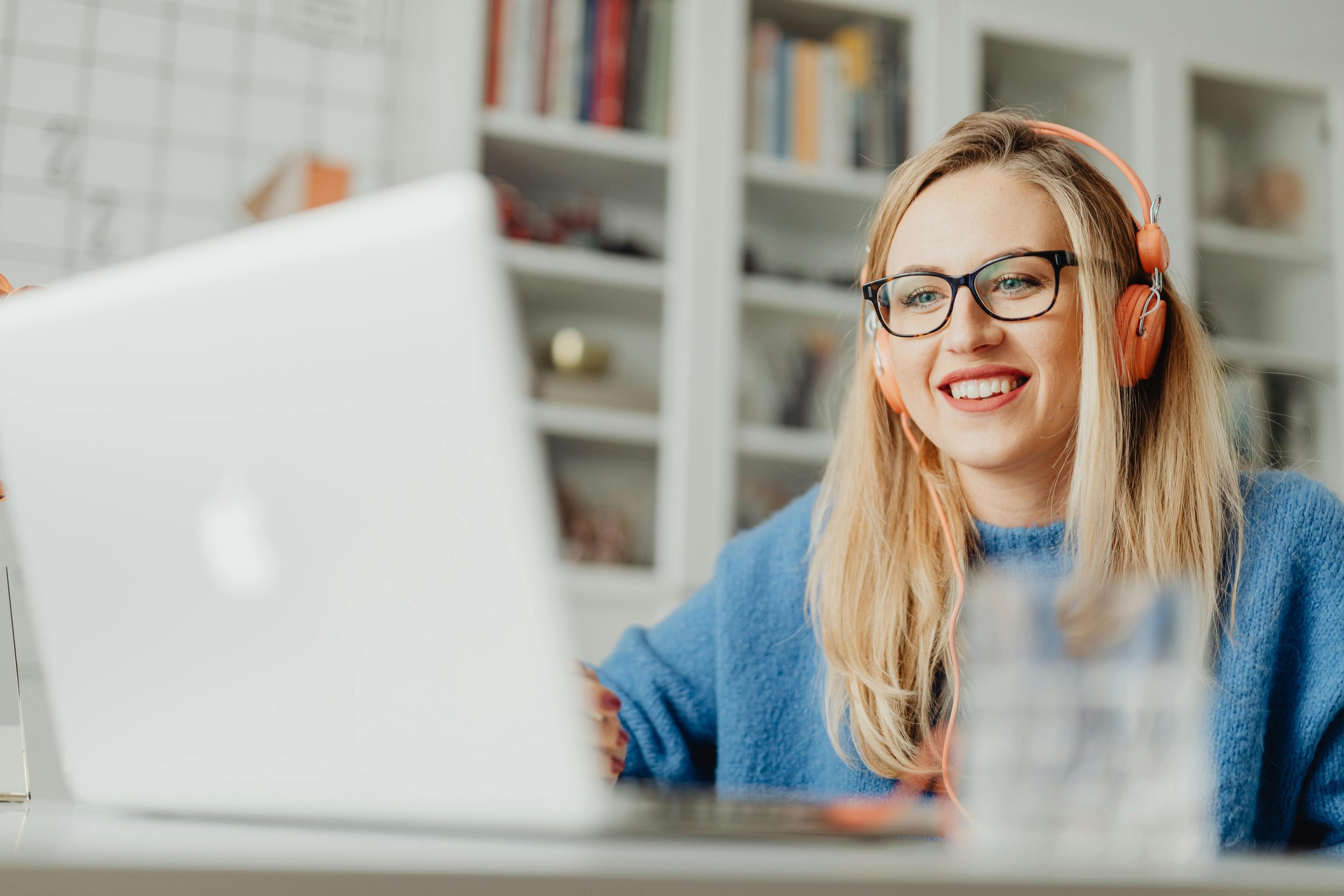 A smiling woman with glasses and blonde hair wearing headphones, working on a laptop in a bright room with shelves in the background.