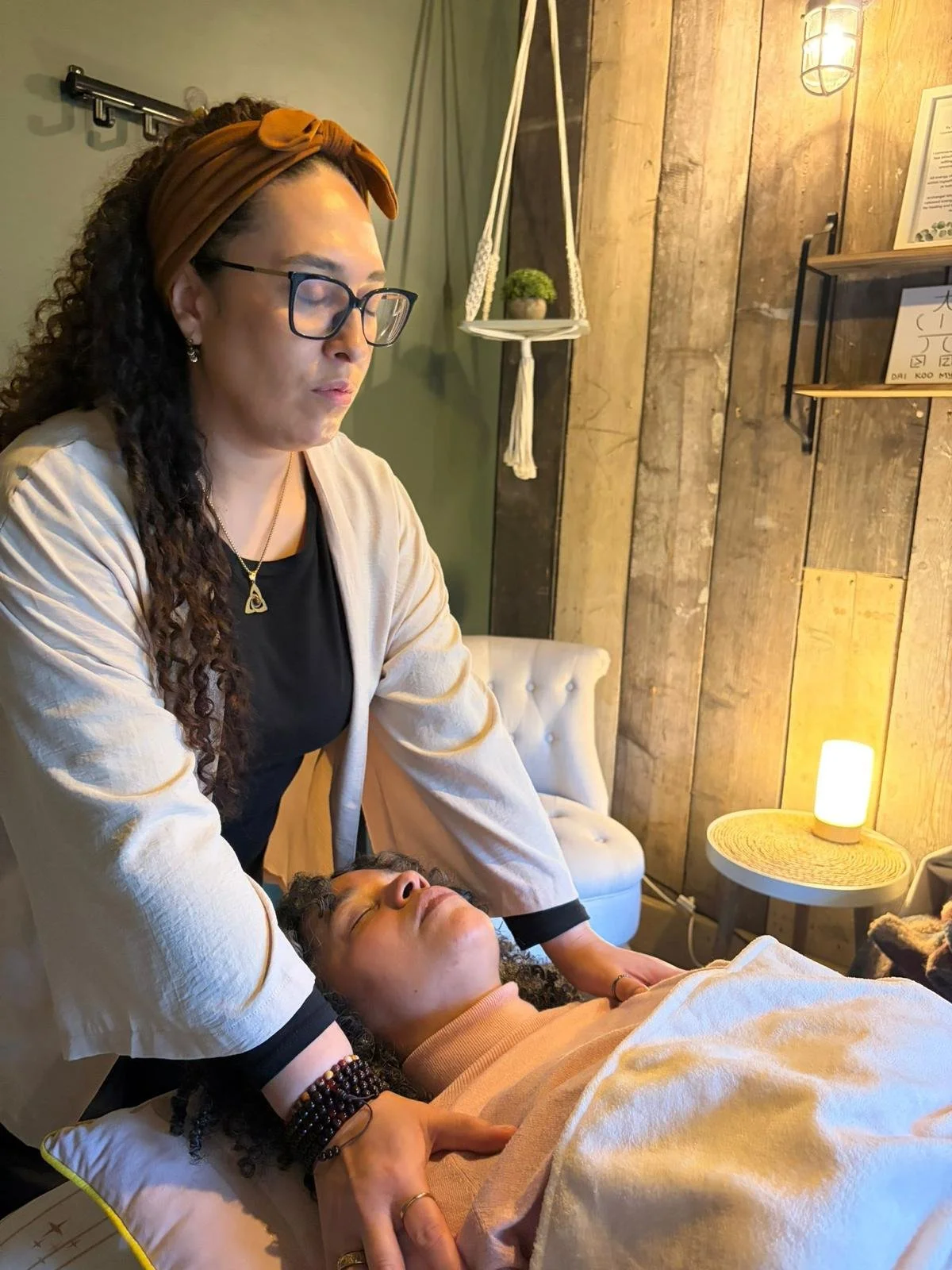 A woman receiving a gentle massage on her forehead from another person, lying on a black pillow with a geometric design, near a large window with a view of trees outside.