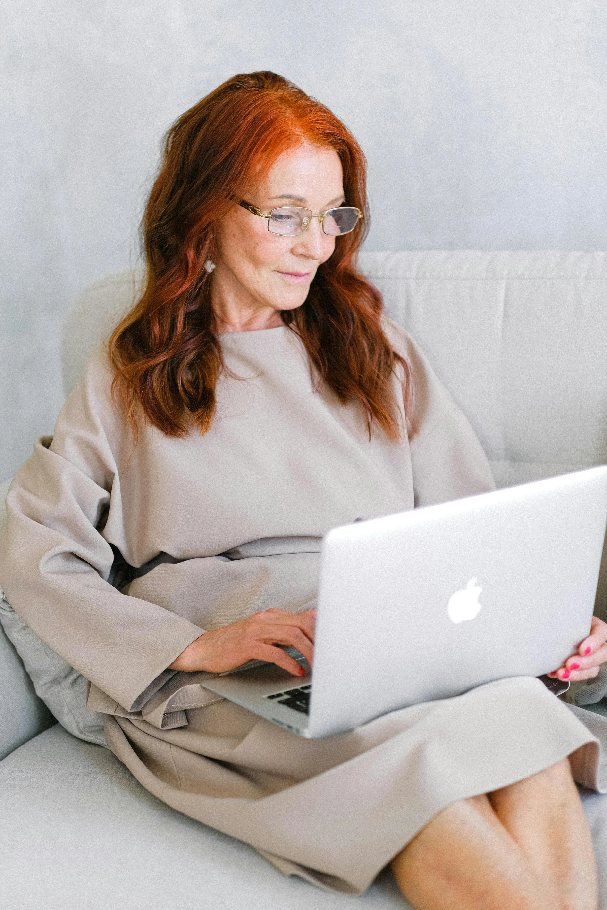 Young woman in a pink hijab sitting on a white couch, writing in a notebook with a laptop on a small round table in front of her, in a bright room with white walls, a floor lamp, and minimal decor.