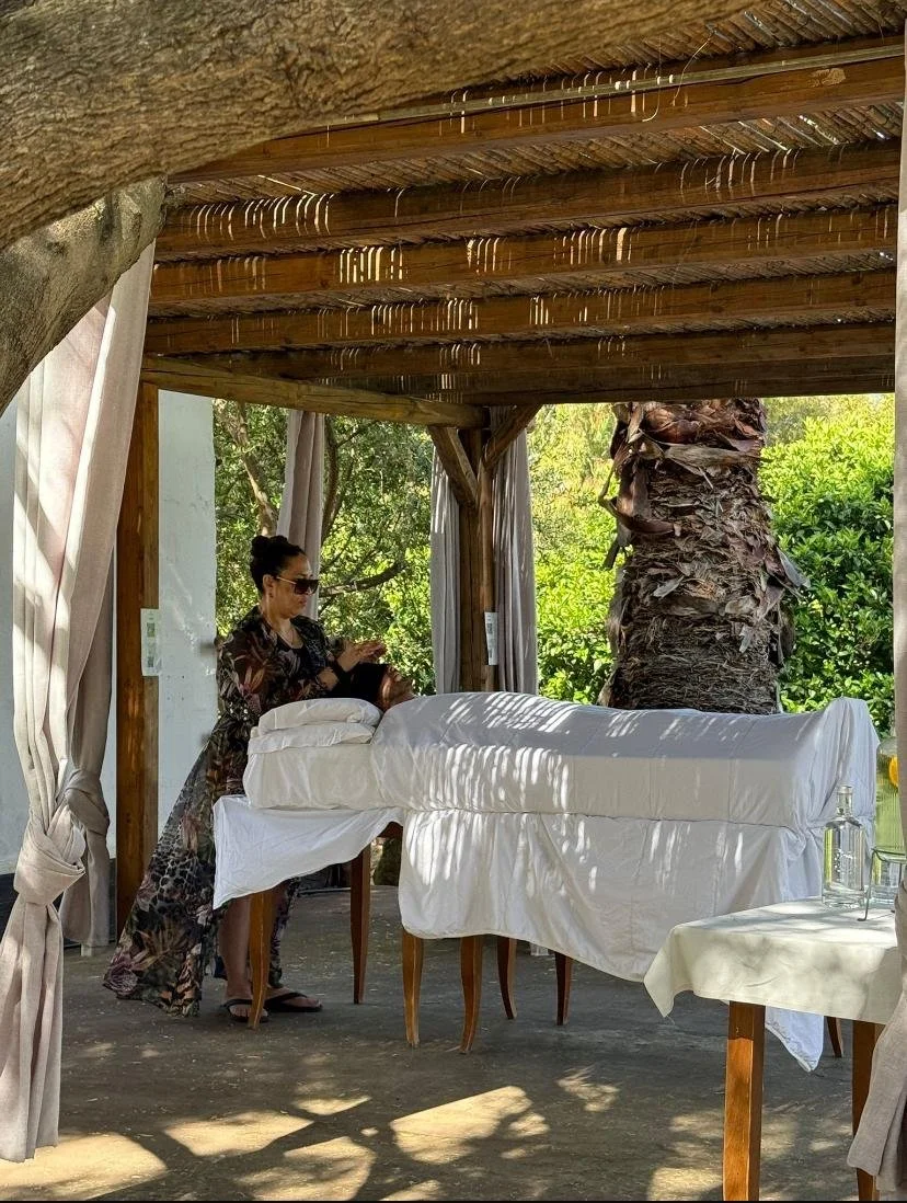 A woman with curly hair lying on her back with eyes closed, while another woman holds a burning incense stick above her face, with smoke rising, in a cozy room.
