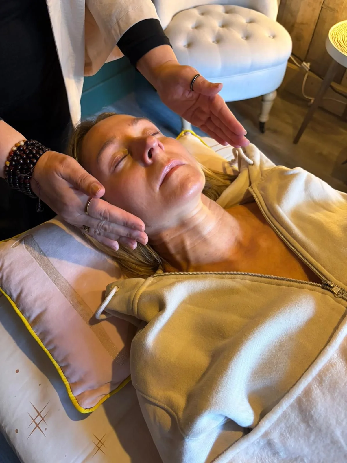 A woman with curly hair lying on her back with eyes closed, while another woman holds a burning incense stick above her face, with smoke rising, in a cozy room.