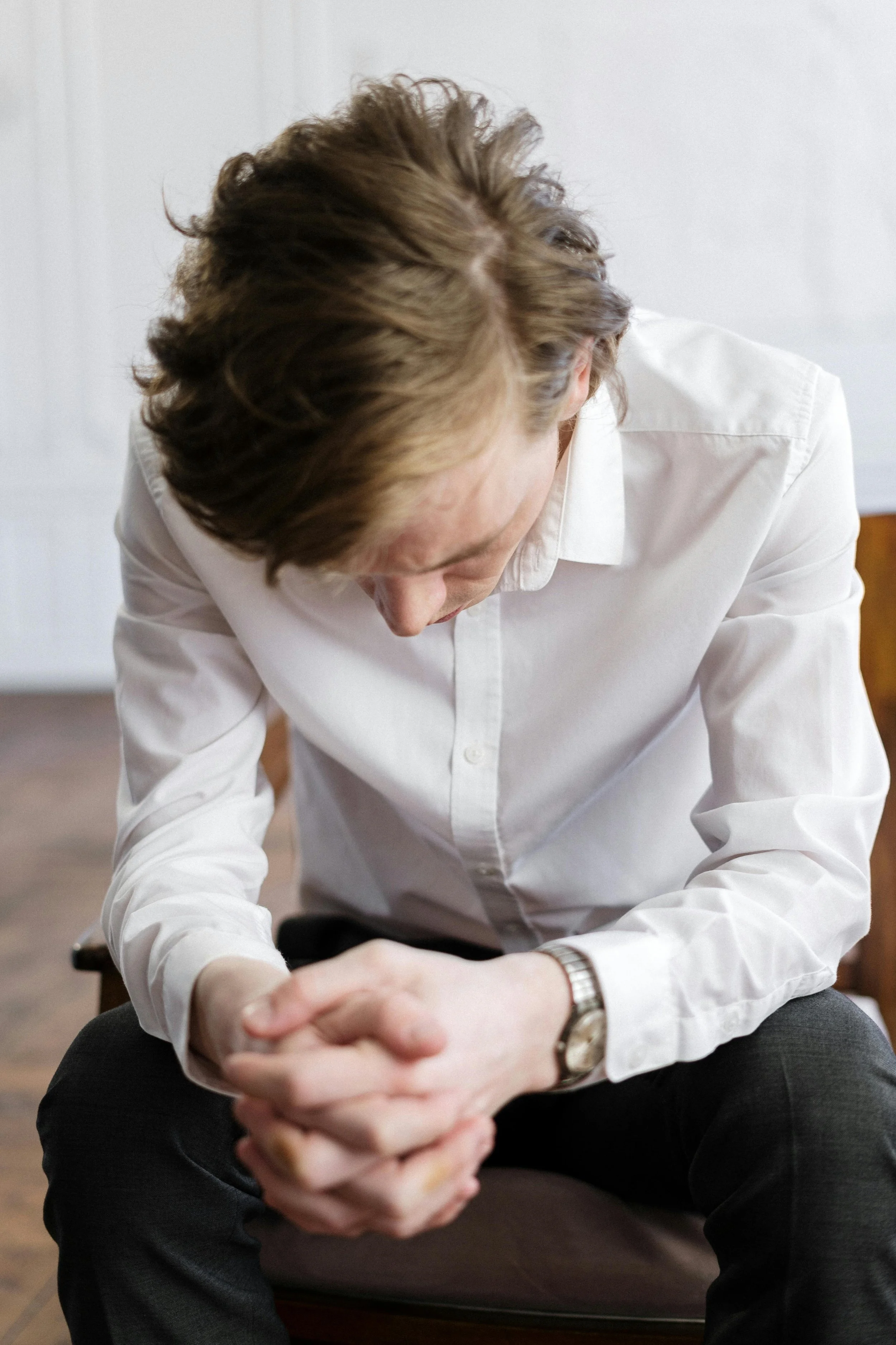 A man with brown hair wearing a white shirt and a wristwatch, sitting with his hands clasped together, head bowed, and looking down.
