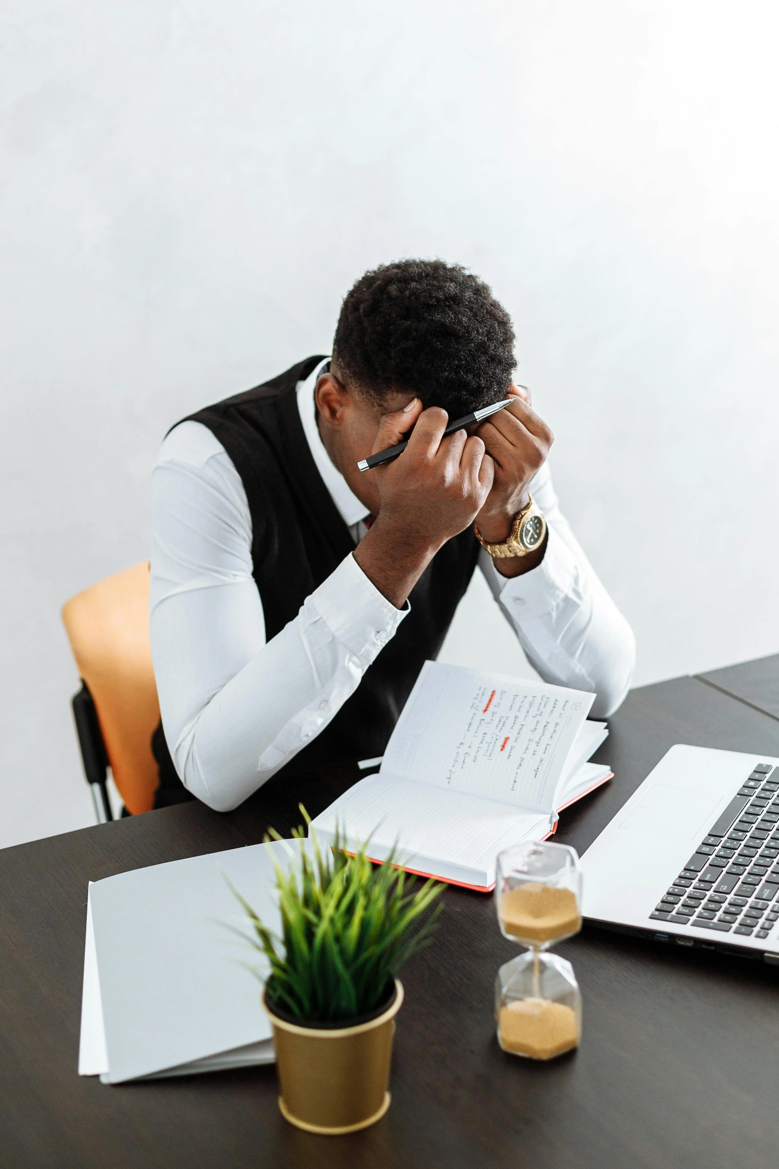 A man sitting at a desk holding his head with one hand and a pen in the other, with an open notebook, a laptop, a small potted plant, a glass hourglass, and a folder on the desk.