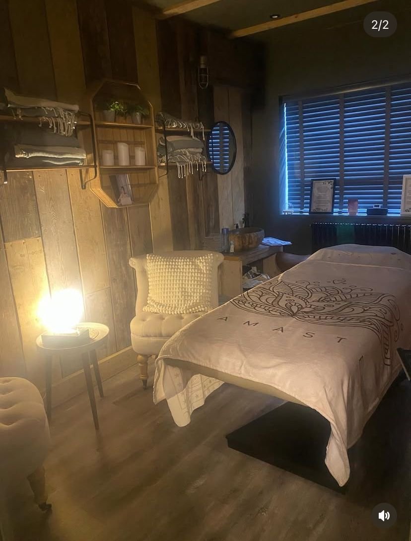 A woman with curly gray hair is lying down with her eyes closed, receiving a Tibetan sound healing session with tuned bowls held by another woman with curly brown hair, standing by a window showing a cloudy sky and green landscape outside.