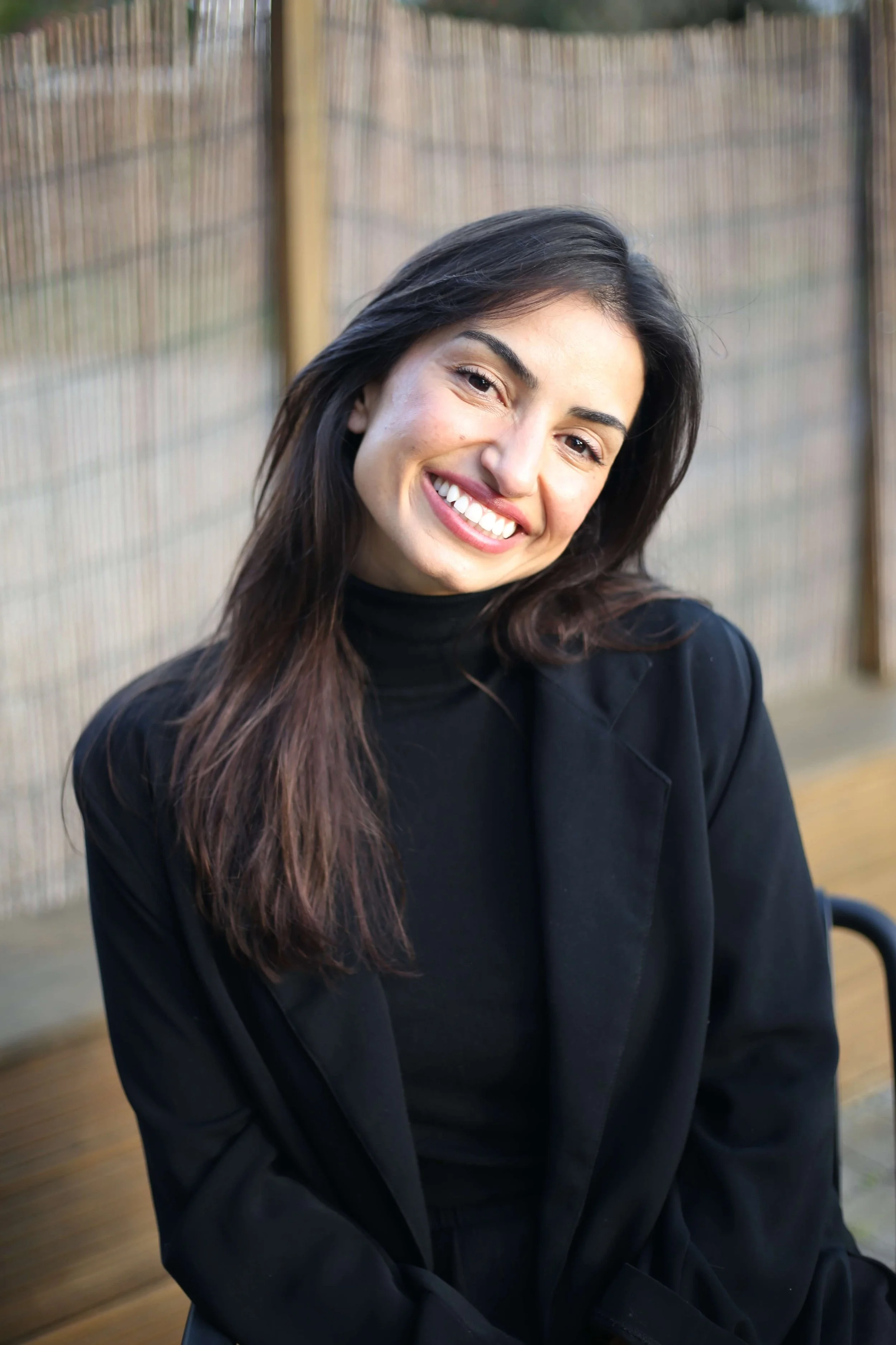 Smiling woman with long dark hair wearing a black blazer and black turtleneck sitting on a wooden bench outdoors with a blurred fence background.