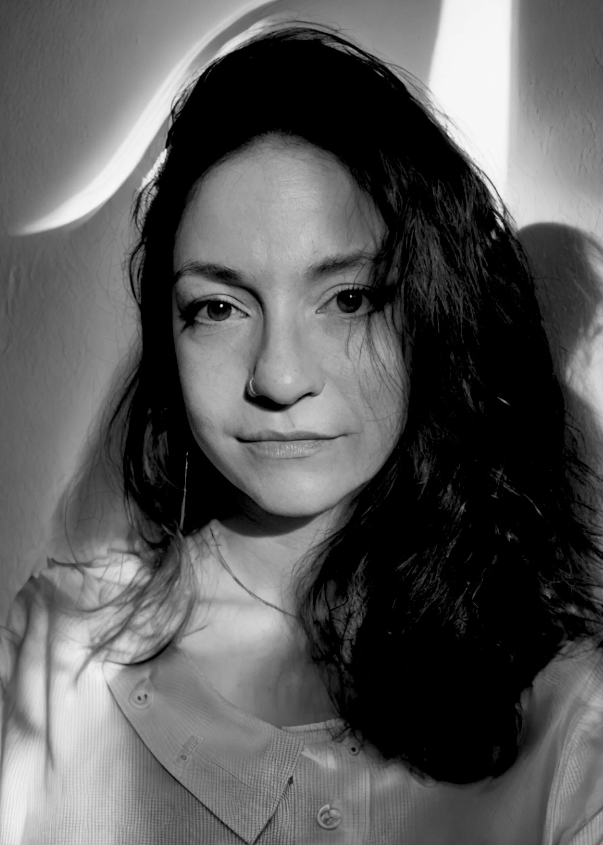 Black and white portrait of a woman with long, wavy dark hair and a nose piercing, wearing a button-up shirt, with strong shadow lighting on her face.