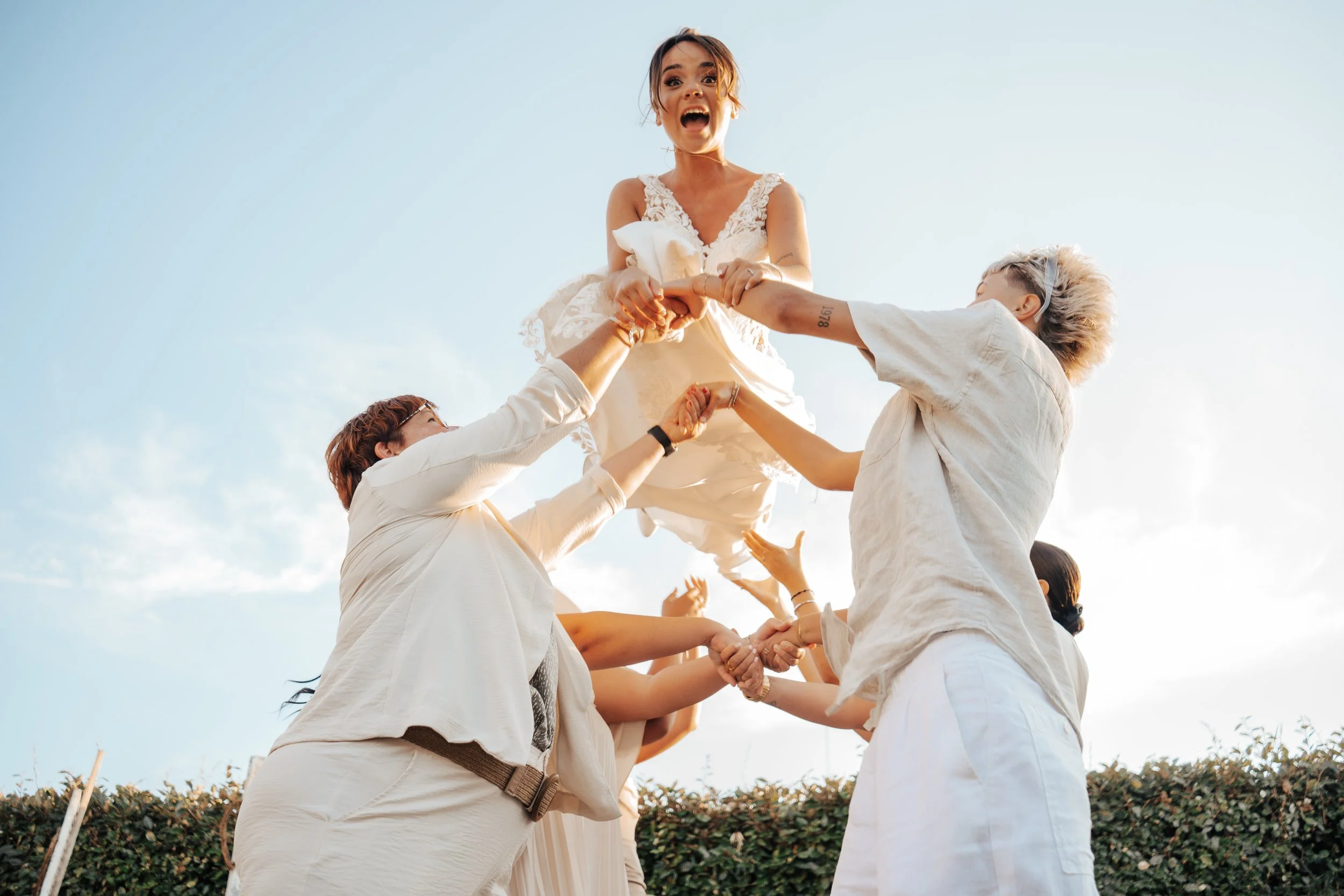 Un groupe de personnes levé haut en l'air soutenant une femme portant une robe blanche, lors d'une célébration extérieure par temps clair.