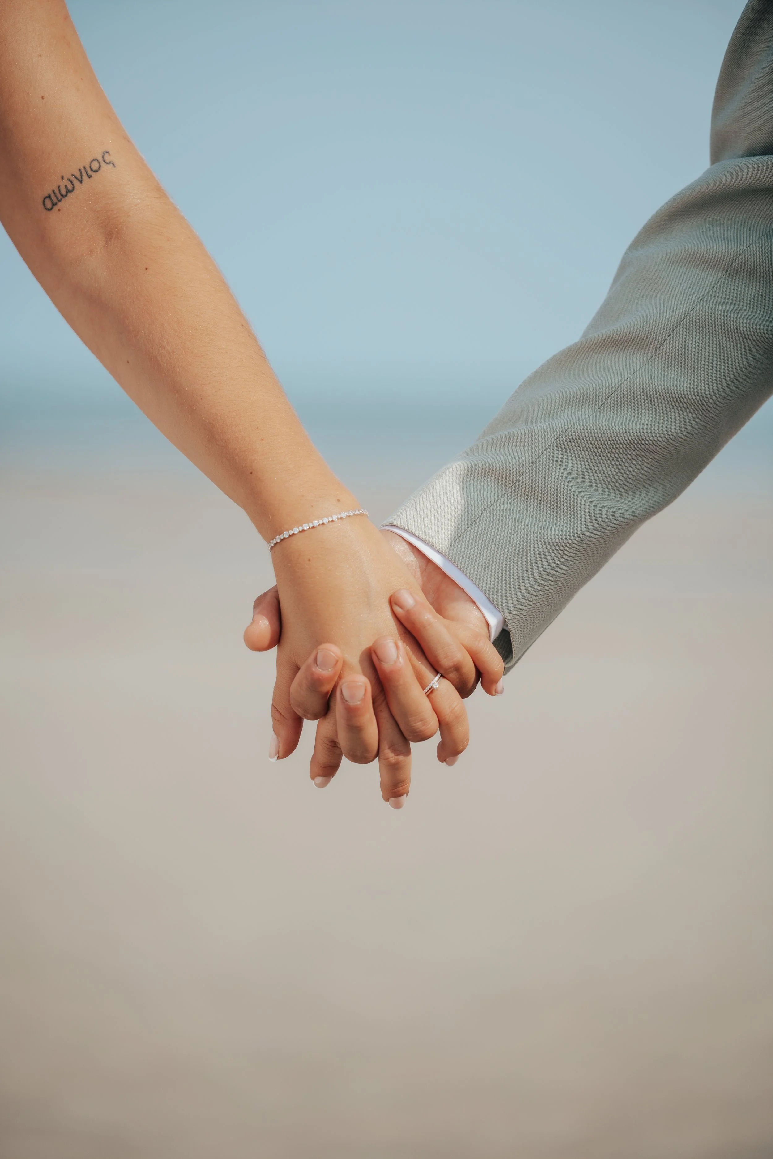 Main tenant la main d'un homme en costume gris, tenant la main d'une femme avec un tatouage et un bracelet, lors d'une cérémonie de mariage sur la plage.