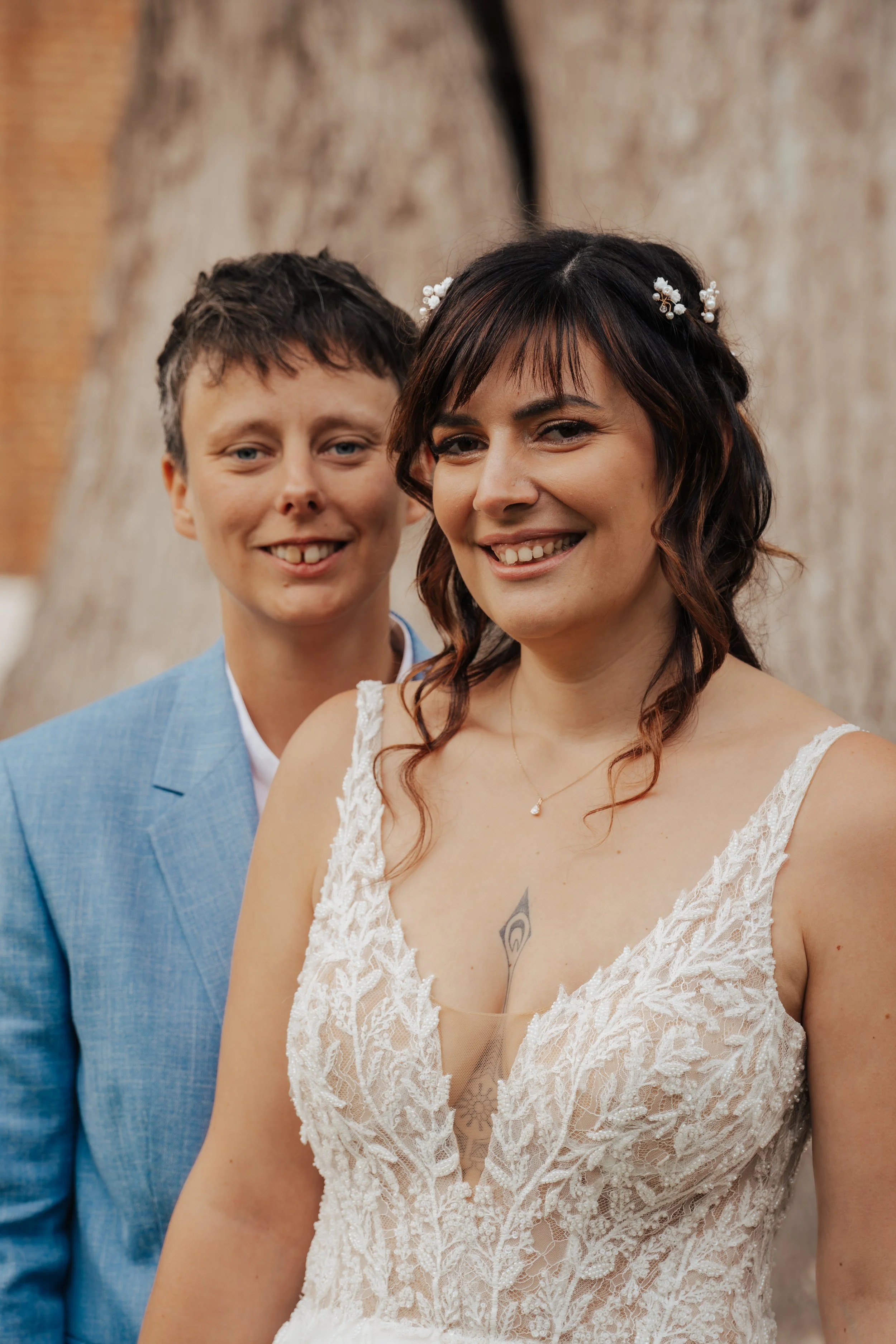 Une femme en robe de mariage avec un tatouage visible sur la poitrine, accompagnée d'un homme en costume bleu, tous deux souriants, devant un arbre.