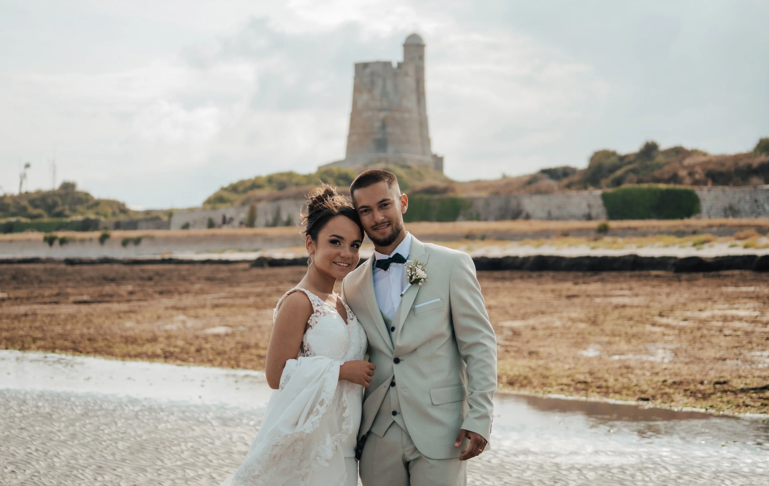 Un couple de mariés souriants en costume blanc se tenant la tête contre la tête sur une plage avec un château en arrière-plan.