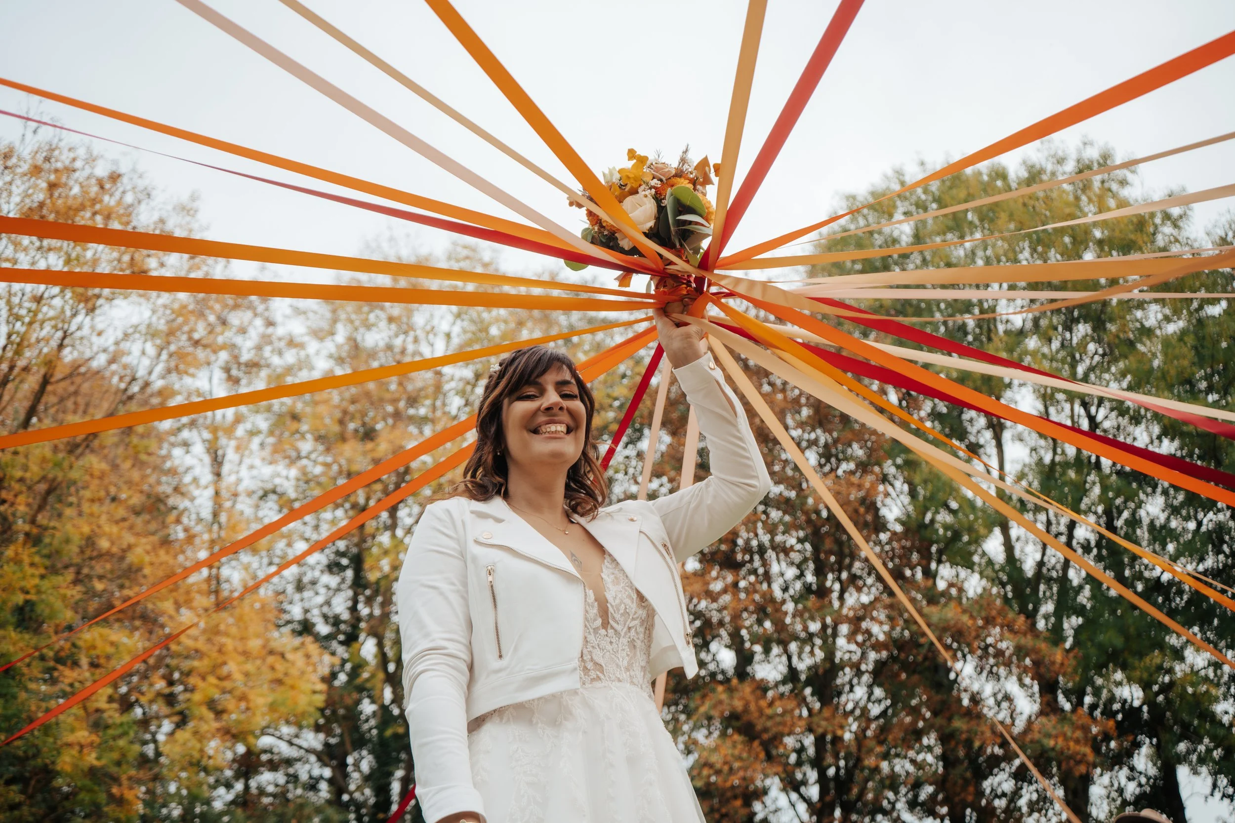Une femme souriante tenant un bouquet, debout sous une toile d'ombrage avec des rubans orange, dans un parc aux arbres aux feuilles d'automne.
