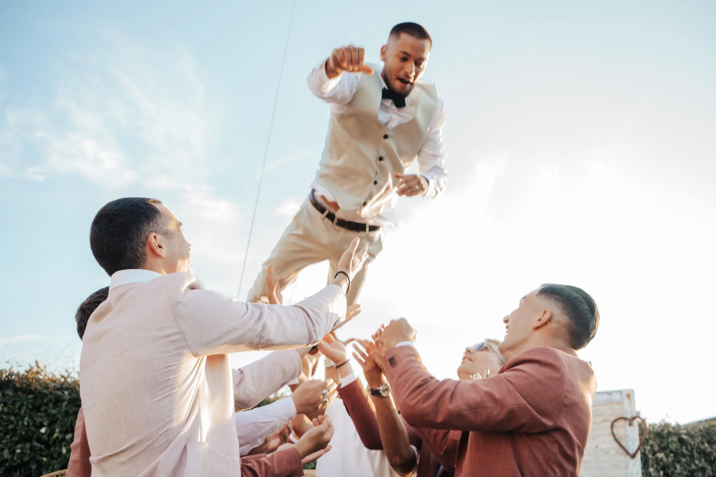 Groupe de personnes en costume, faisant une étape de soutien pour un homme en costume blanc, qui semble danser ou faire une acrobatie lors d'une célébration en plein air.