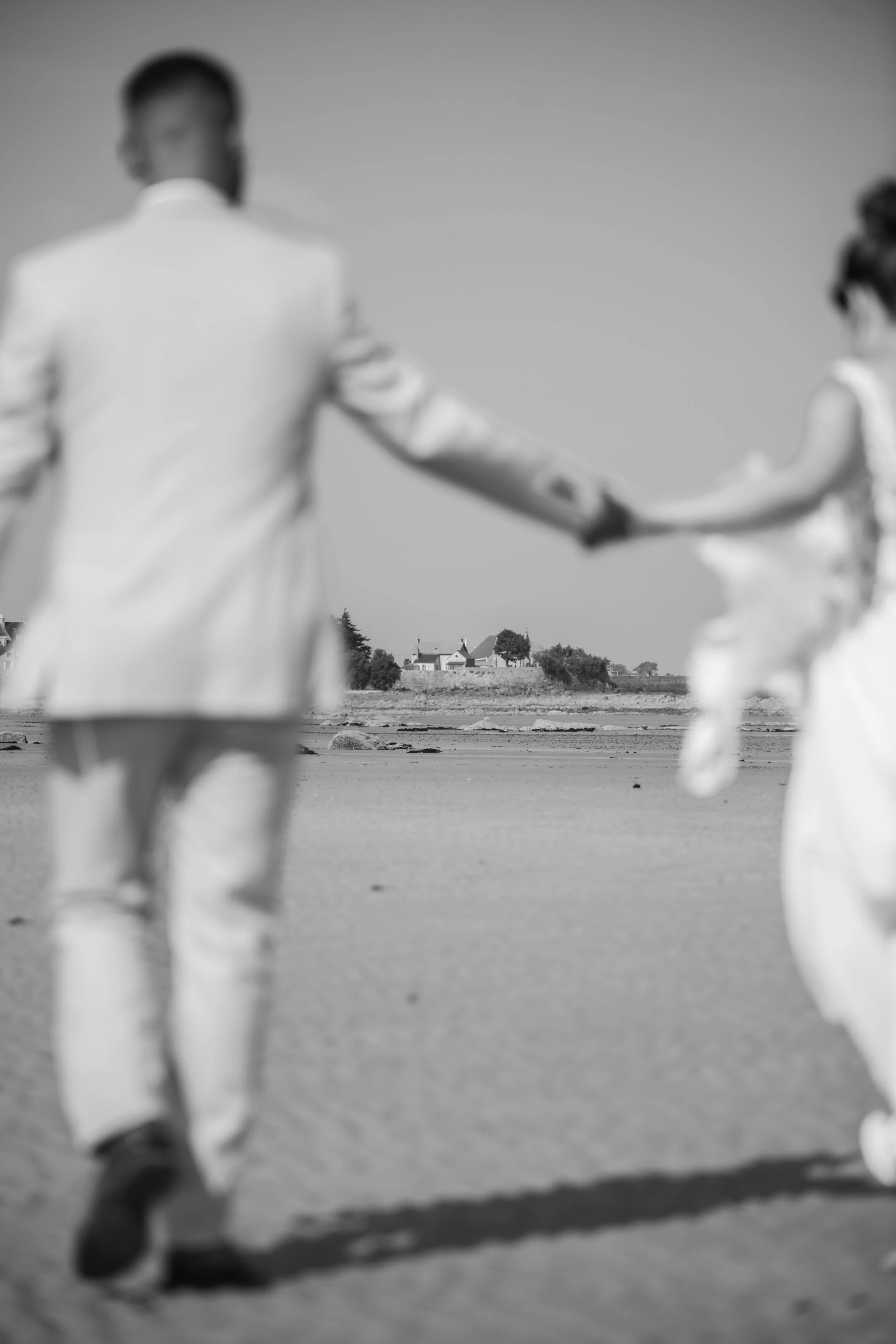 Une photo en noir et blanc d'un couple marchant main dans la main sur une plage, avec des maisons en arrière-plan.