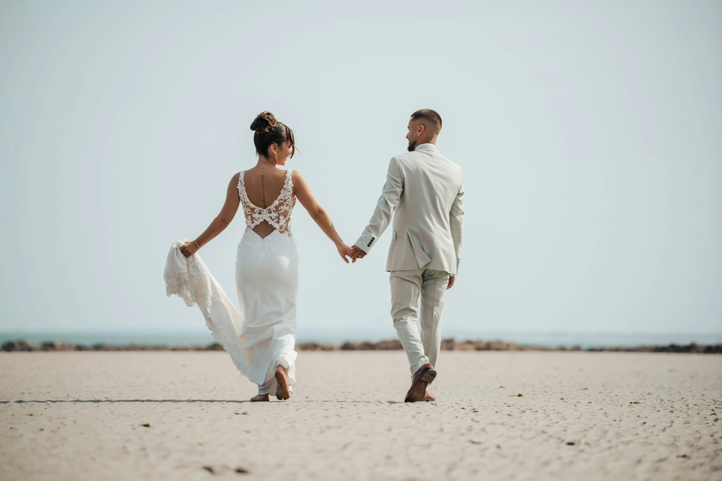 Un couple en vêtements de mariage marche sur une plage, tenant la main, avec la mer en arrière-plan.
