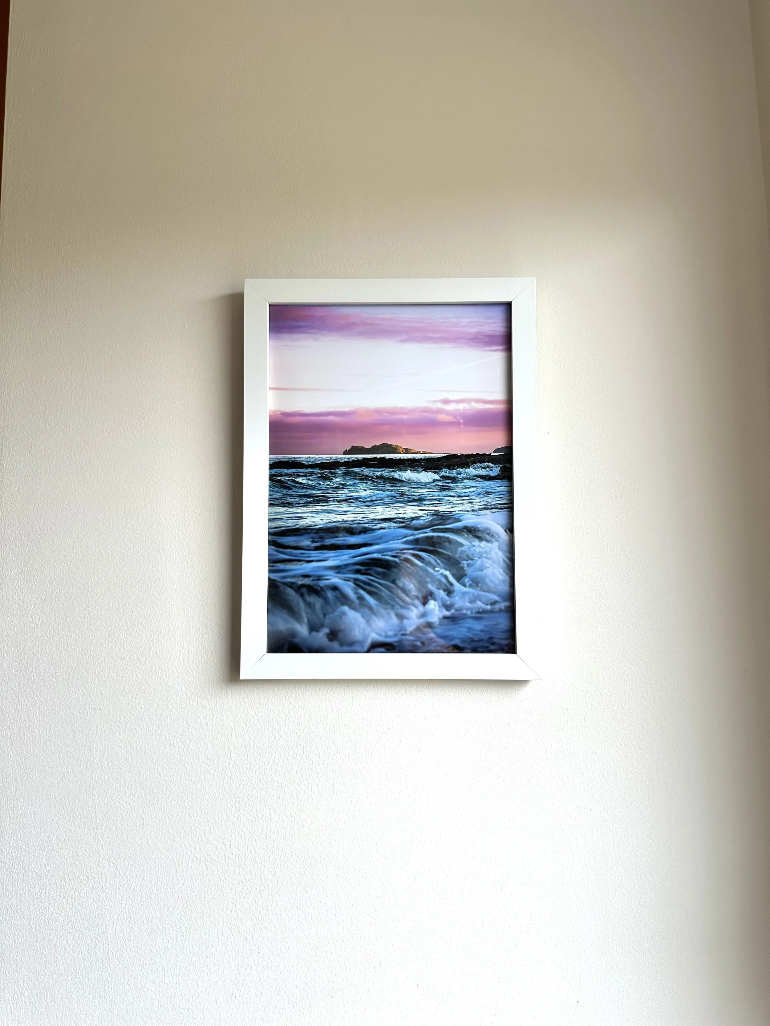 Framed photograph of ocean waves crashing against rocks during sunset with pink and purple sky.