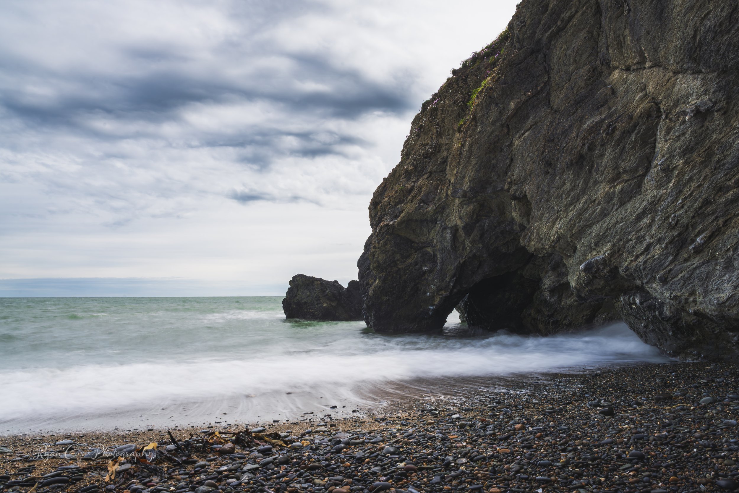 A rocky beach with dark pebbles leading to the ocean, a large rock formation on the right, and a cloudy sky overhead.