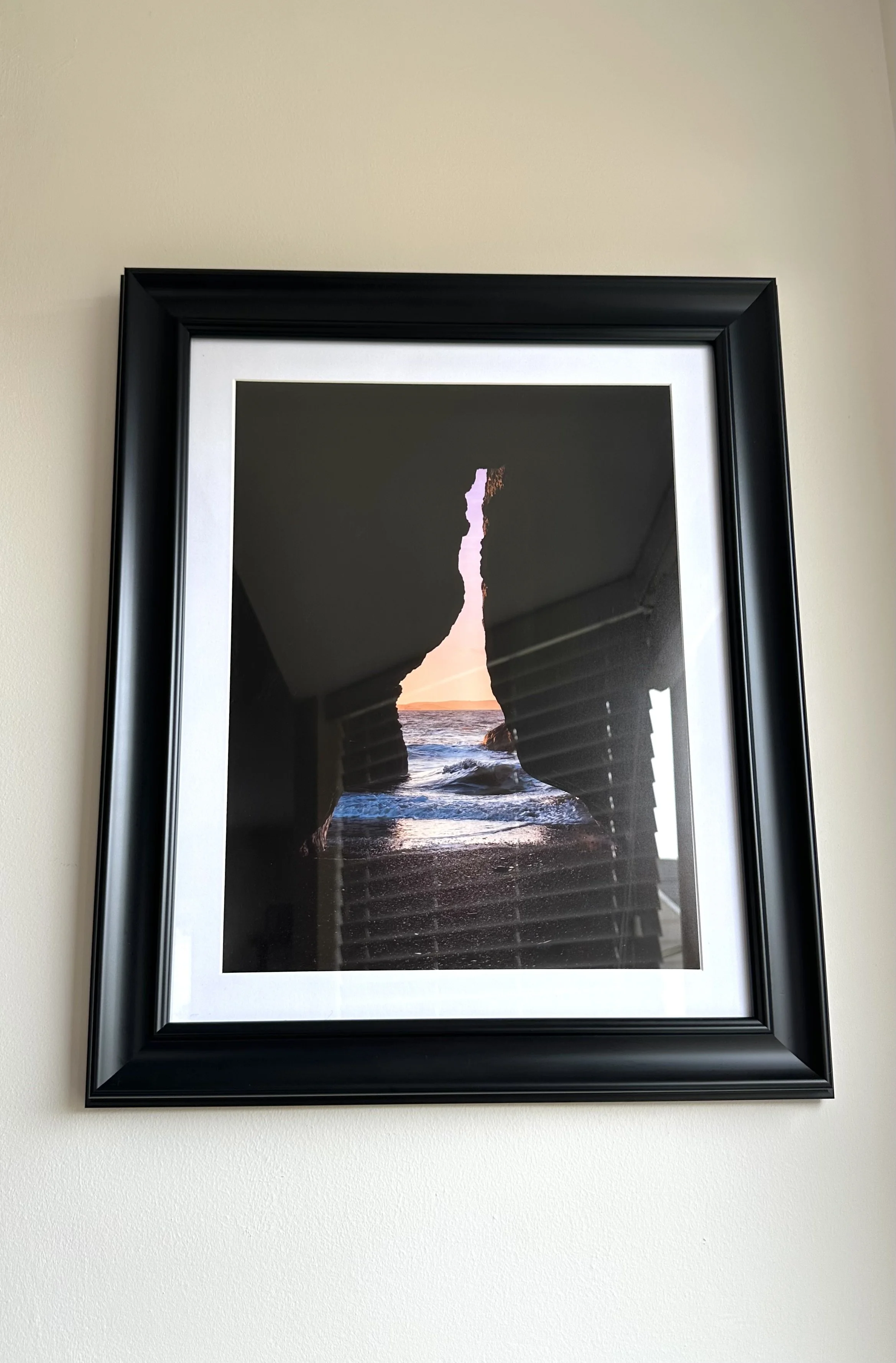 Framed photograph of a rocky seaside cave opening at sunset, with waves crashing inside, hanging on a beige wall.