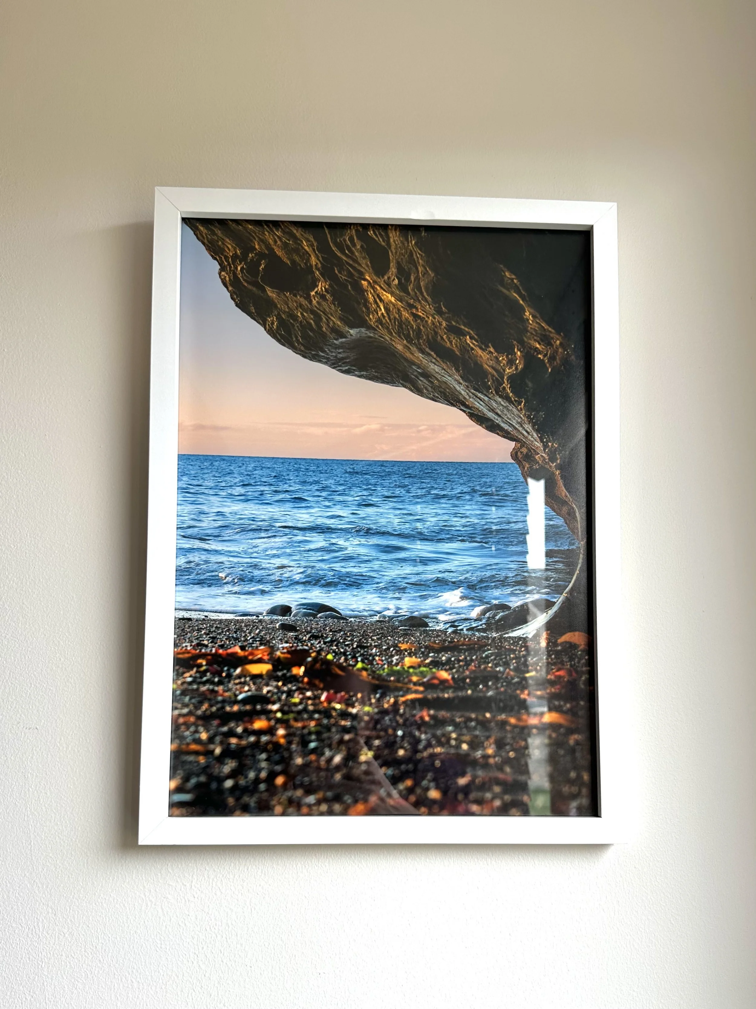 Framed photograph of a rock formation over the ocean at sunset with a pebble beach in the foreground.