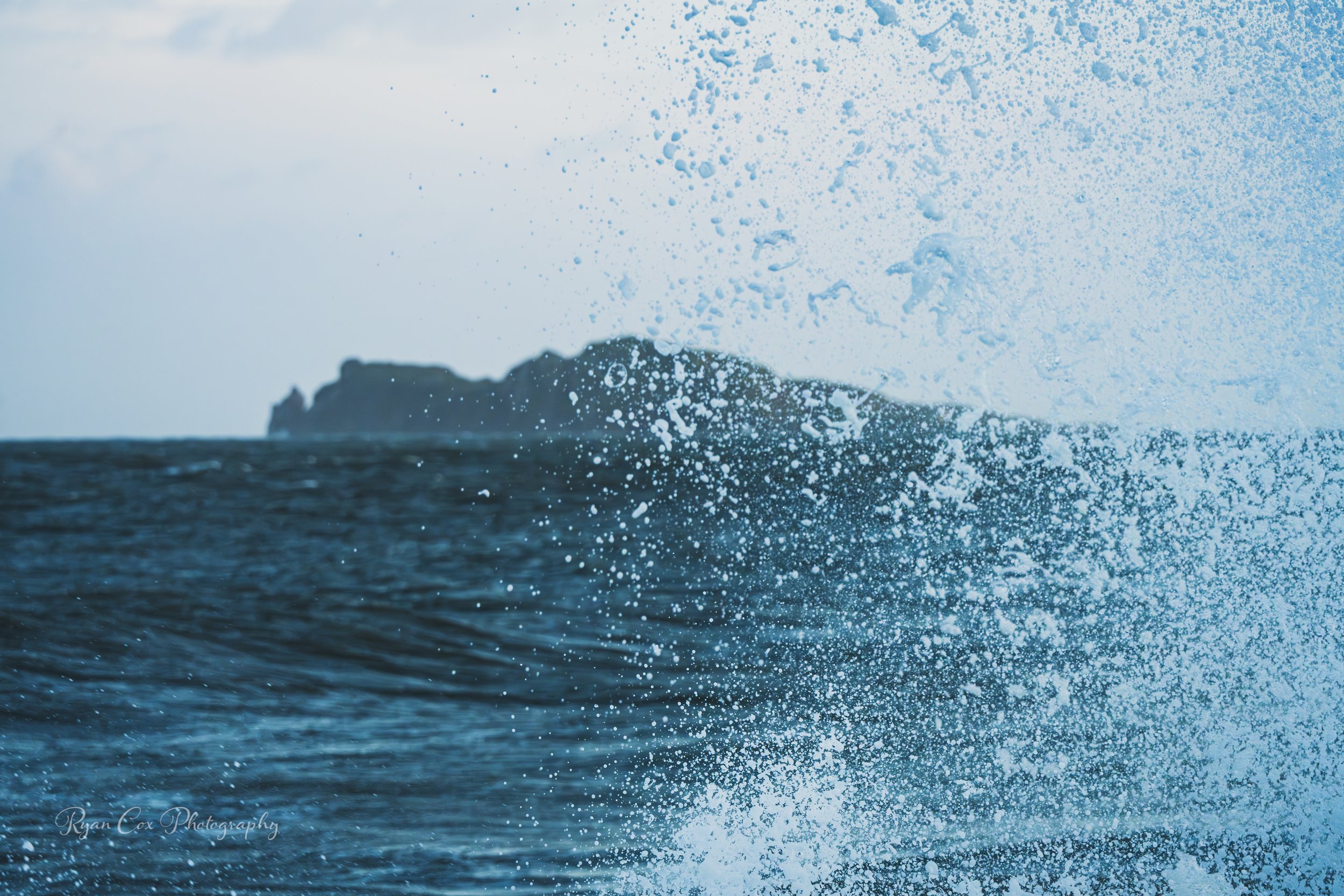 A large ocean wave crashing with spray and water droplets in the air, with a distant rocky island or landmass in the background.