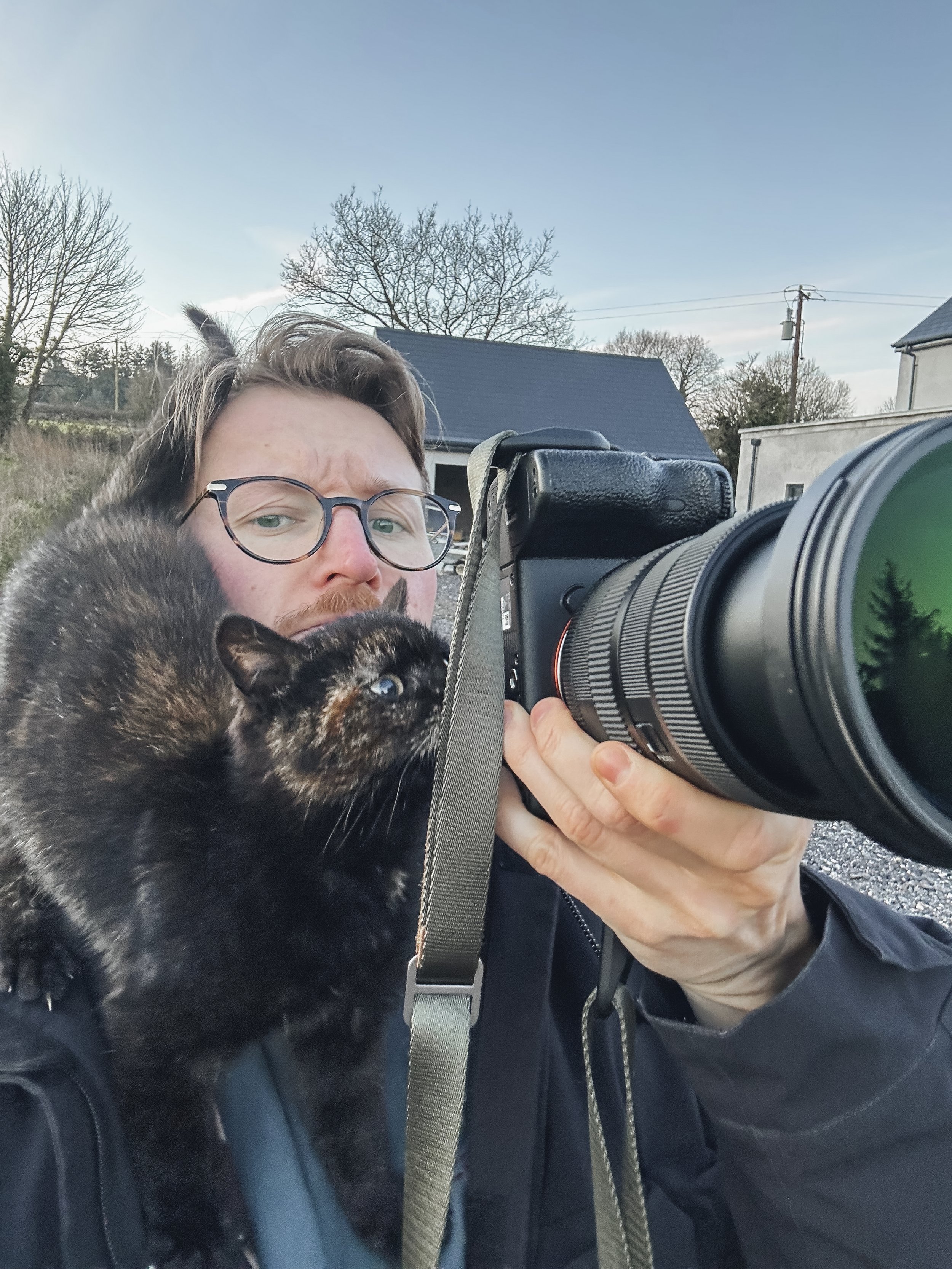 A man with glasses takes a selfie outdoors with a camera while holding a black and brown tortoiseshell cat. The background shows a house, trees, and a clear sky.