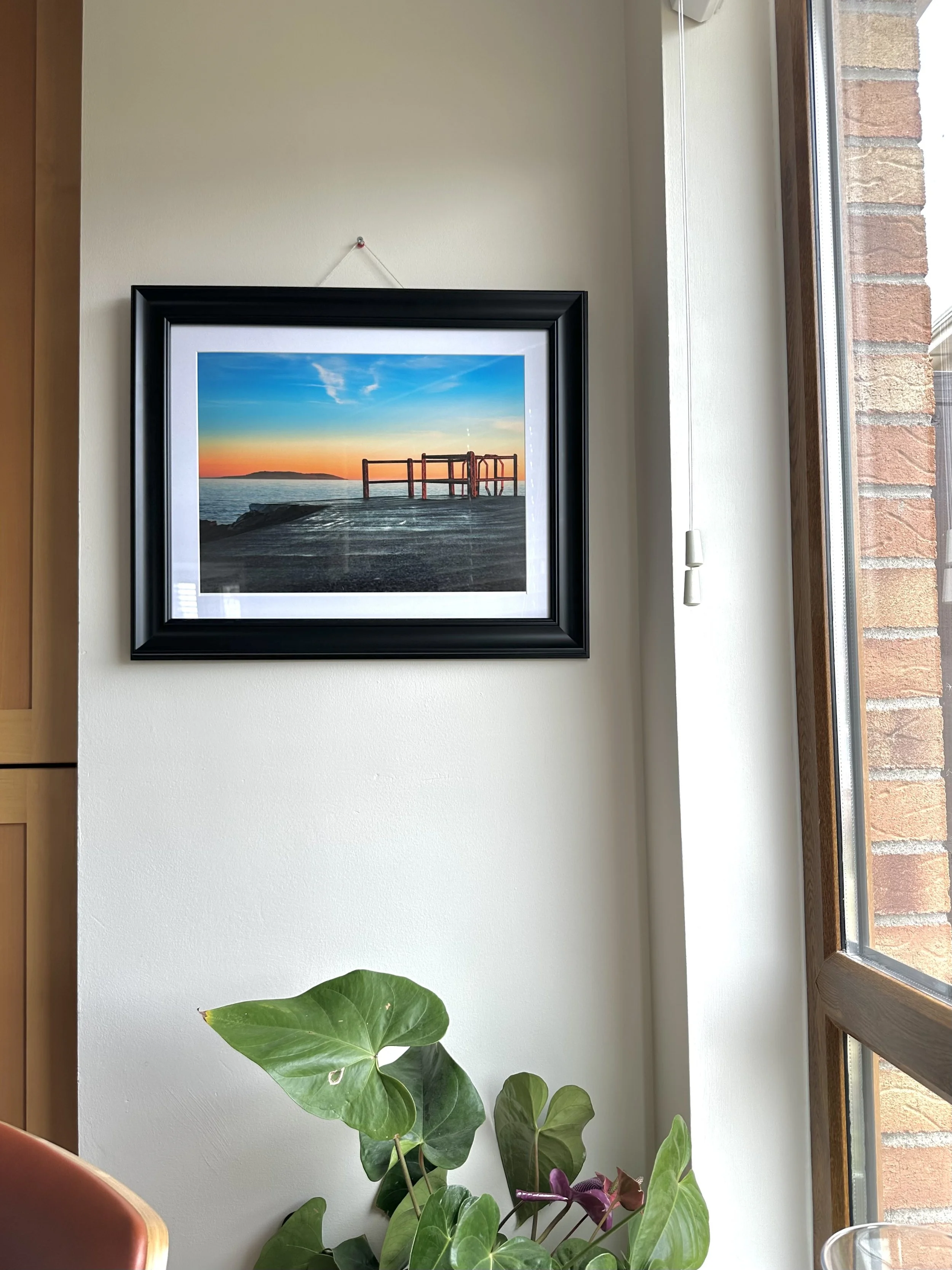 A framed landscape photograph of a sunset over a body of water, hanging on a white wall near a window with a brick wall outside. There are green and purple leafy plants at the bottom of the image.