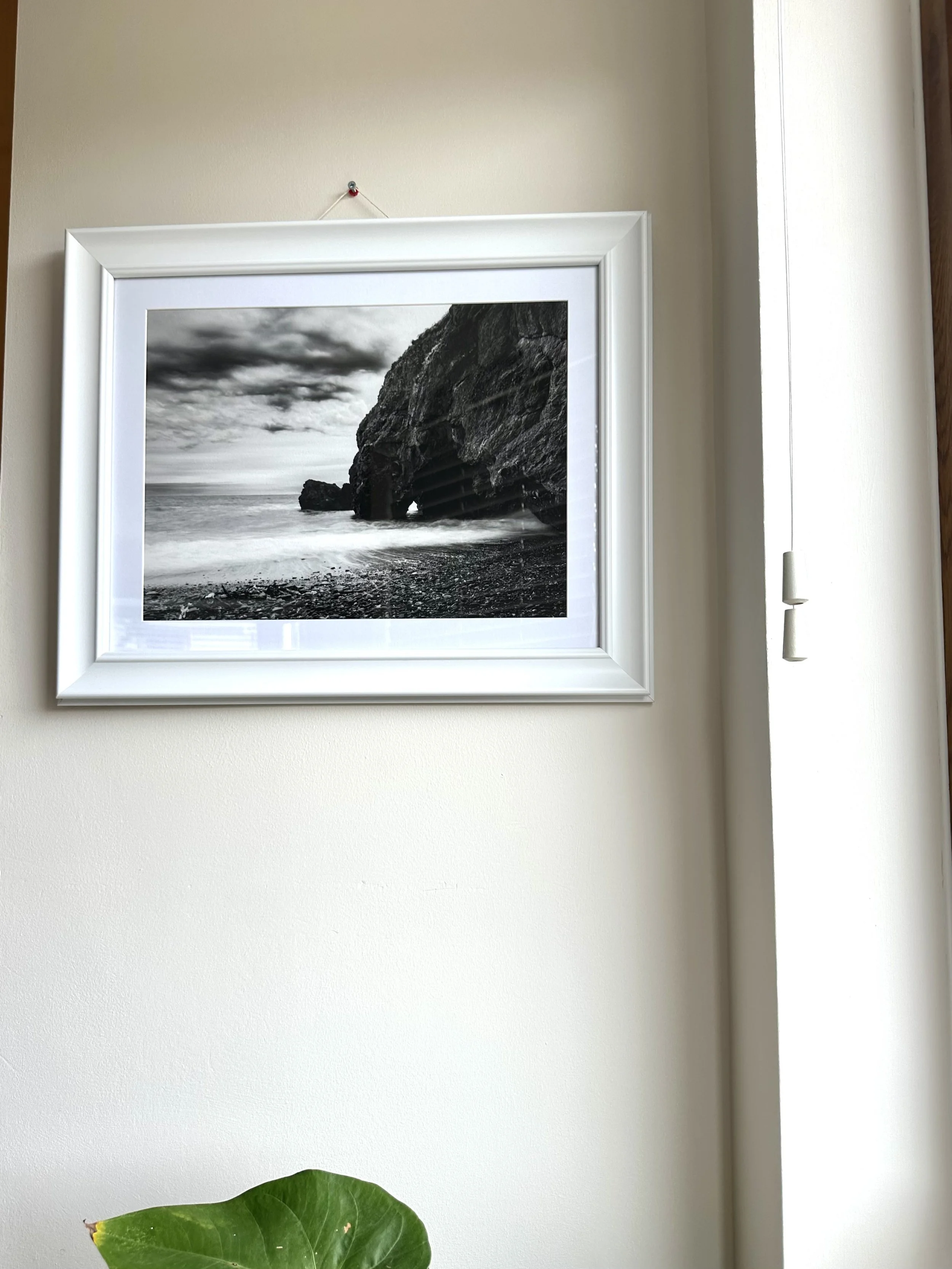 A framed black and white photograph of a rocky coastal scene with a large cliff, ocean waves, stormy sky, hanging on a wall with a white border and hook. Part of a green plant leaf is visible at the bottom.