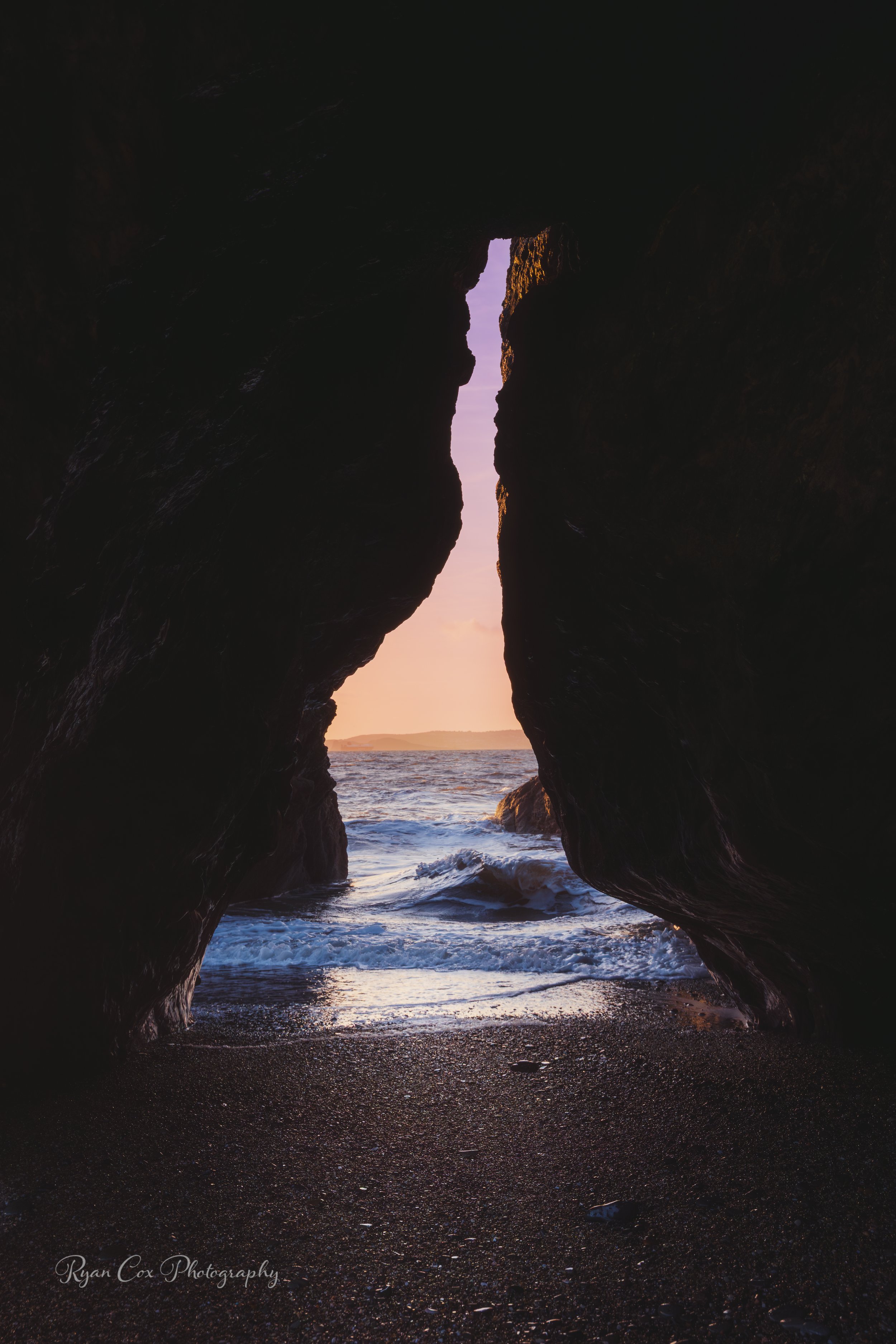 Sunset view through a rocky ocean cave, with waves crashing on the beach and a pinkish sky in the background.
