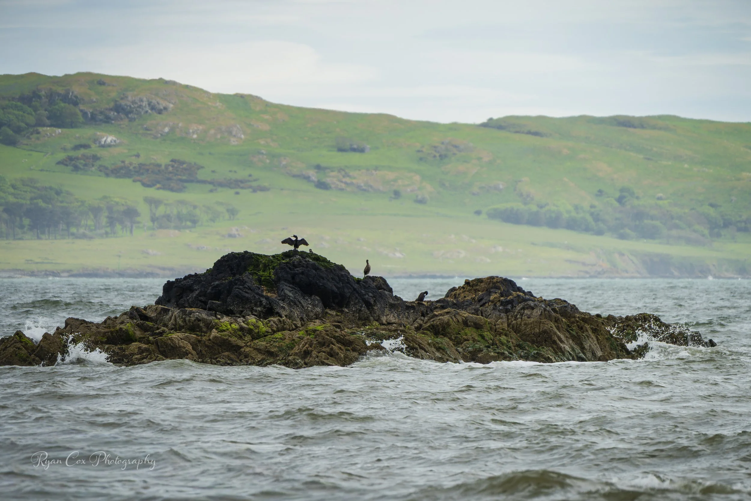 A lighthouse stands on a rocky outcrop in the ocean with waves crashing against the rocks. In the background, green hills stretch across the horizon under a partly cloudy sky.