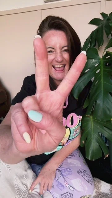 Squarespace web designer who is wearing vibrant pyjamas and is holding her fingers out in front of her in the peace 'V' sign. She is sitting next to a Monstera plant.