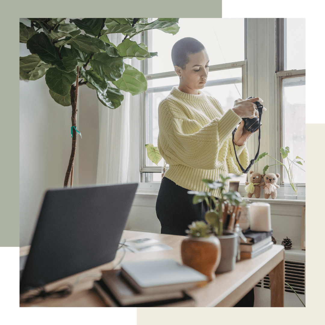 A woman with short hair wearing a yellow sweater looks at a camera while standing near a window decorated with small stuffed animals, with plants and a laptop on the desk.
