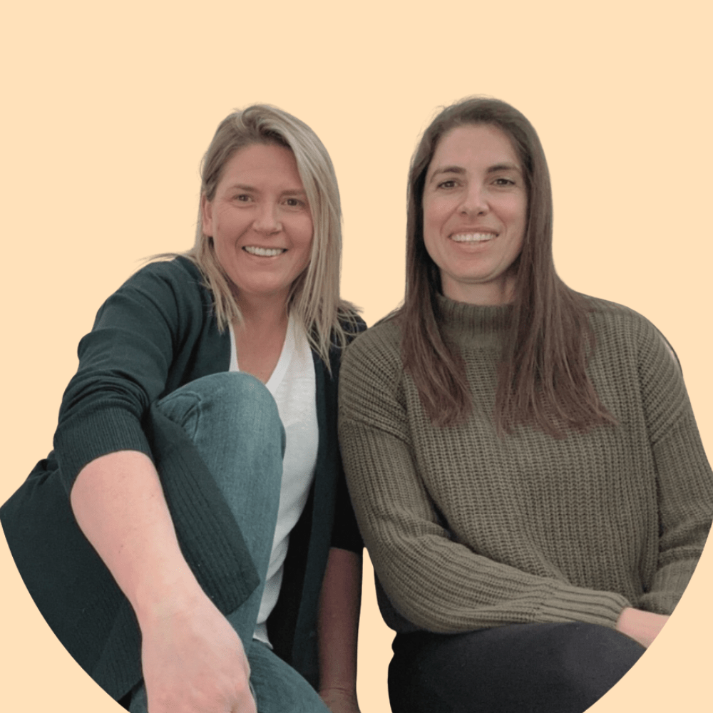 Two women sitting close together and smiling at the camera against a light peach background.