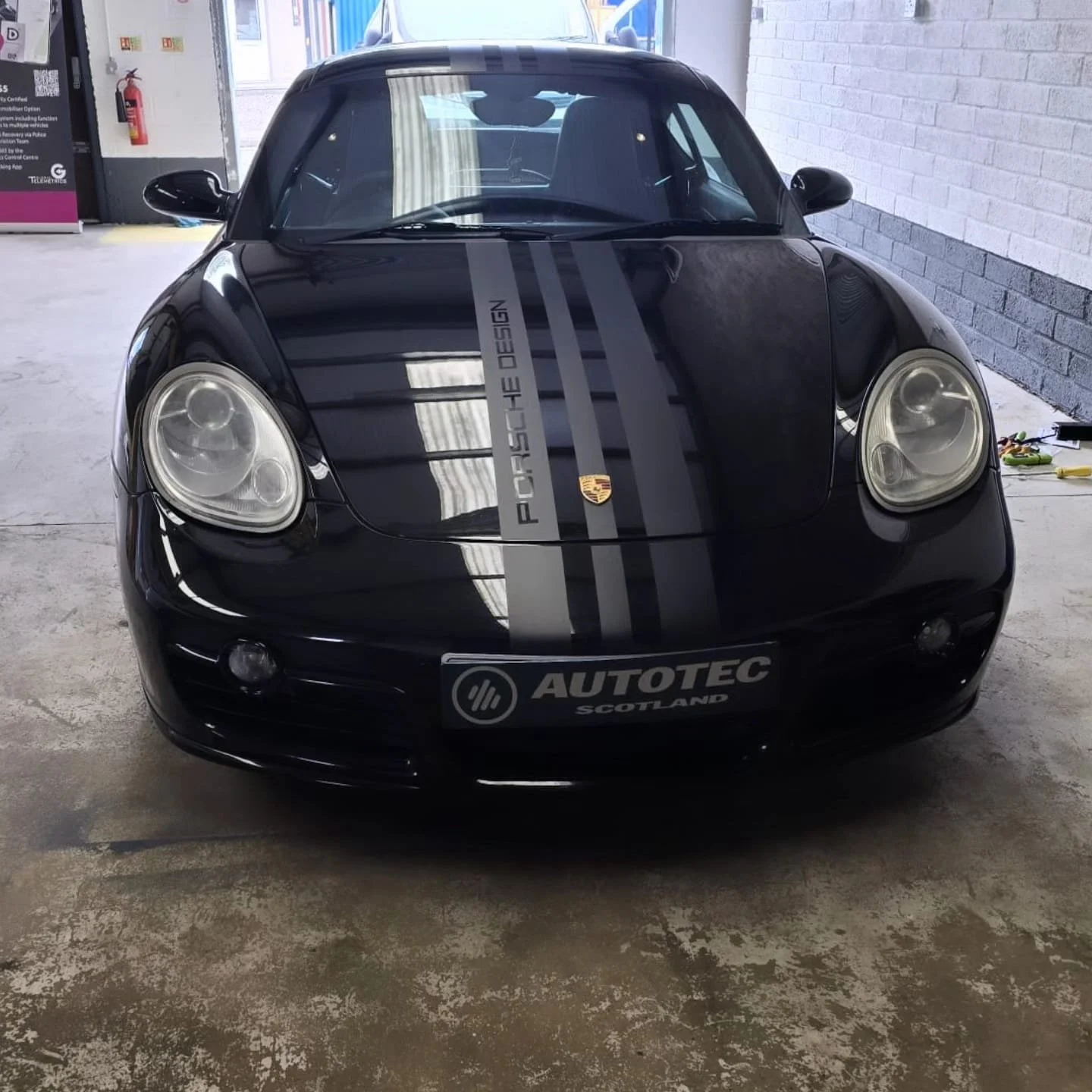 Black Porsche sports car with gray racing stripes featuring Porsche branding on the hood, parked in an indoor garage.