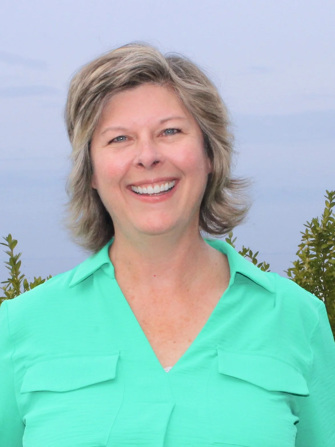 A smiling woman with short, wavy hair wearing a light green shirt standing outdoors with trees and a cloudy sky in the background.