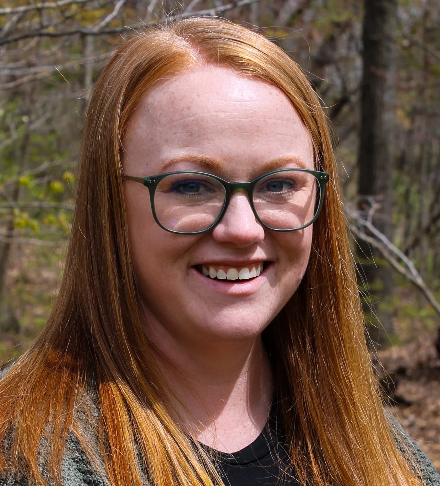 A woman with long red hair, smiling, against a background of trees.