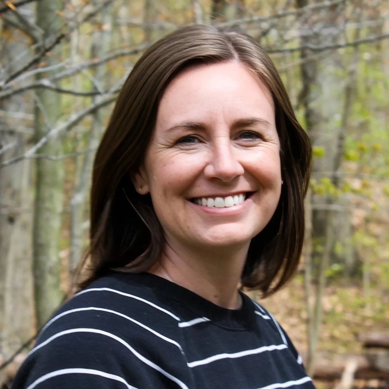 A woman with shoulder length brown hair, smiling, standing in front of trees. 