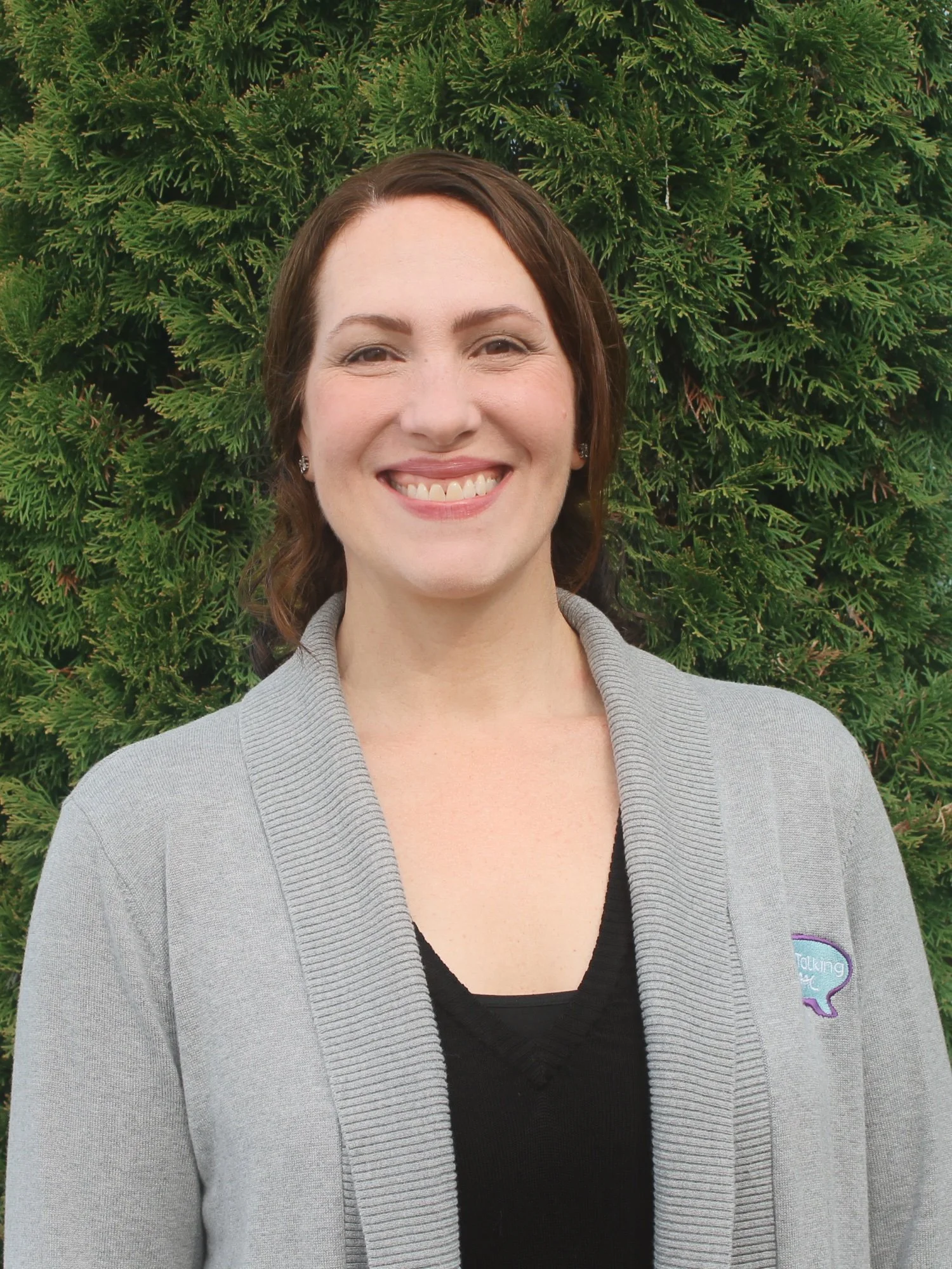 A smiling woman with shoulder-length brown hair, wearing a gray cardigan with a logo on the chest, standing outdoors in front of green shrubbery.