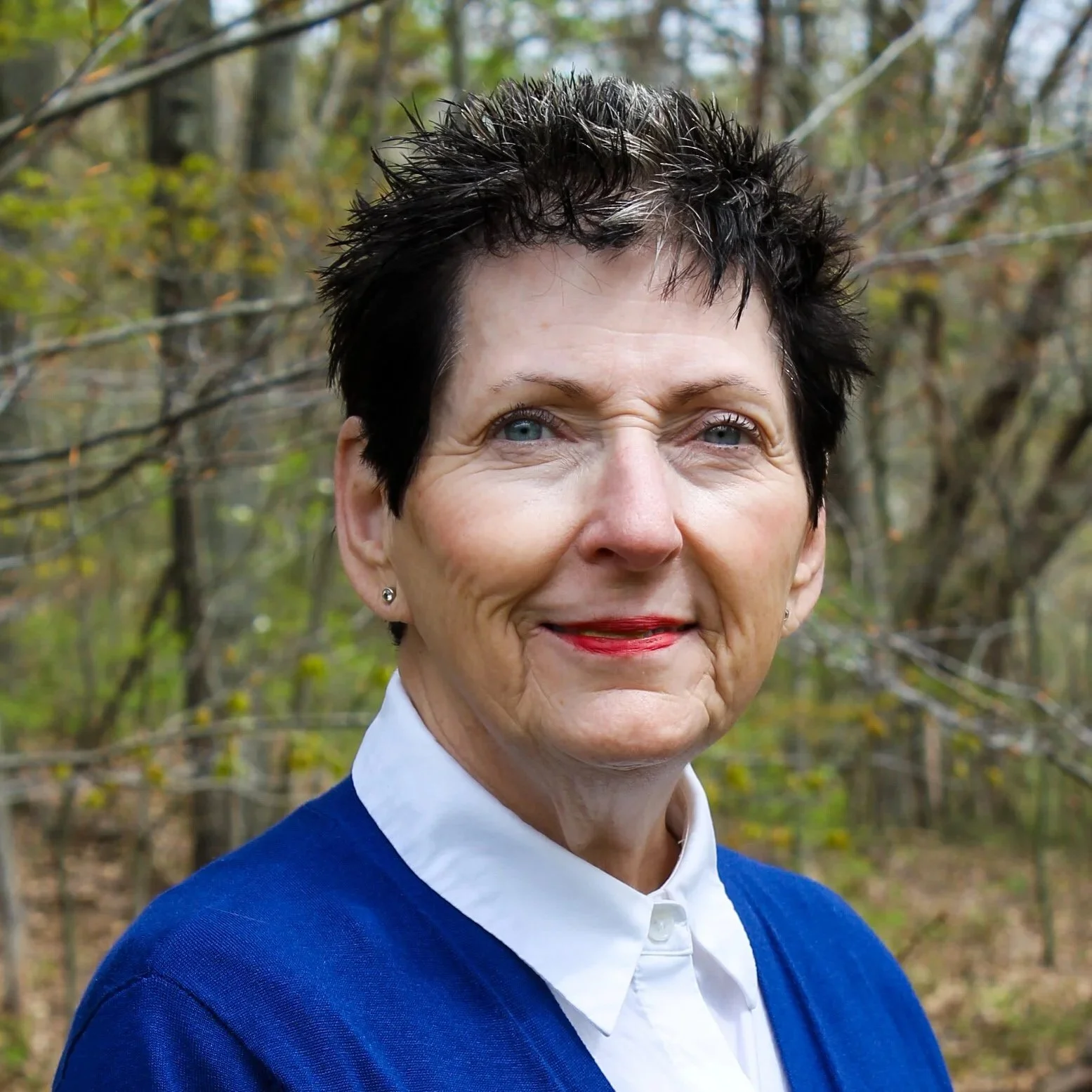 A woman with short dark hair standing outdoors against a background of trees.