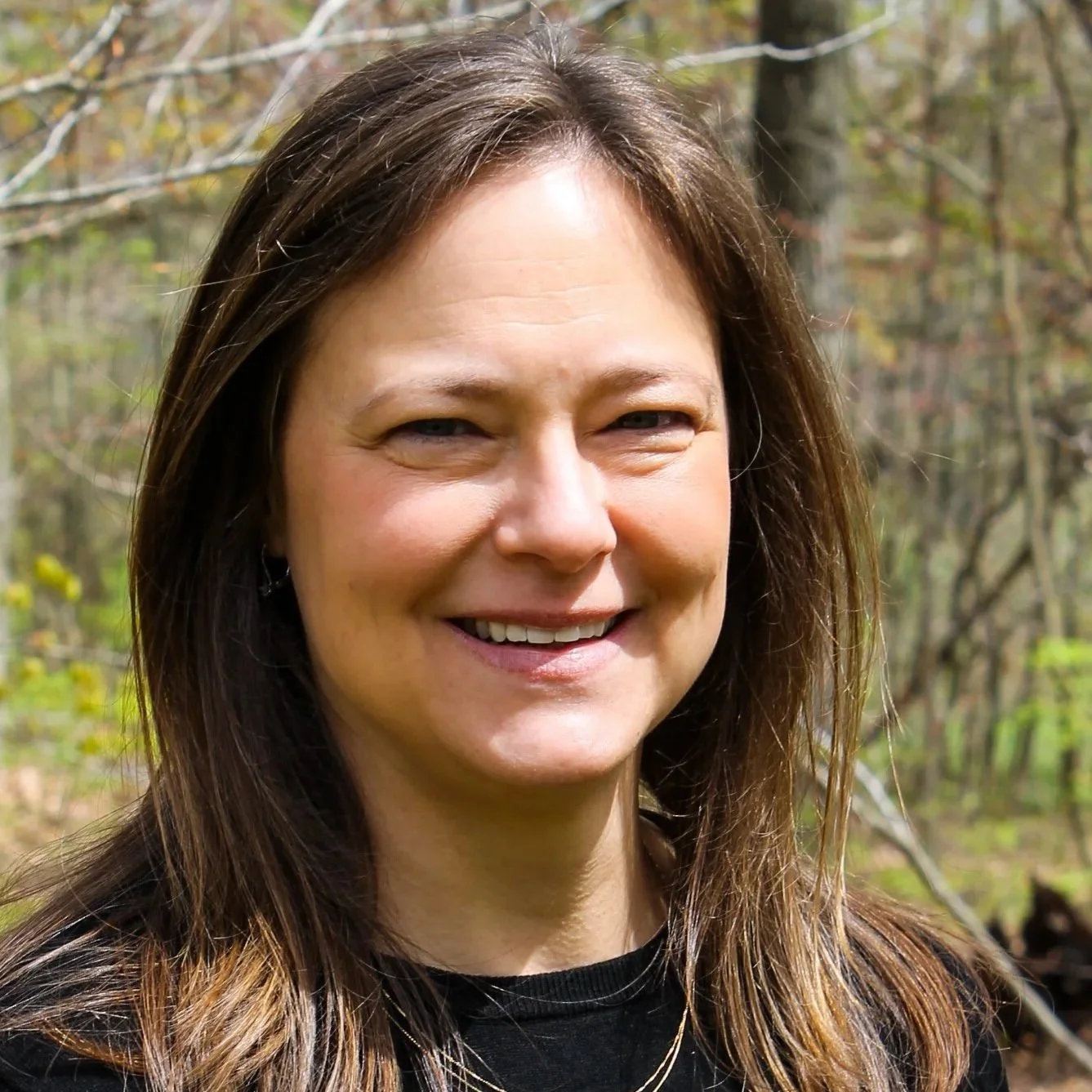 A woman with long brown hair, with trees in the background. 