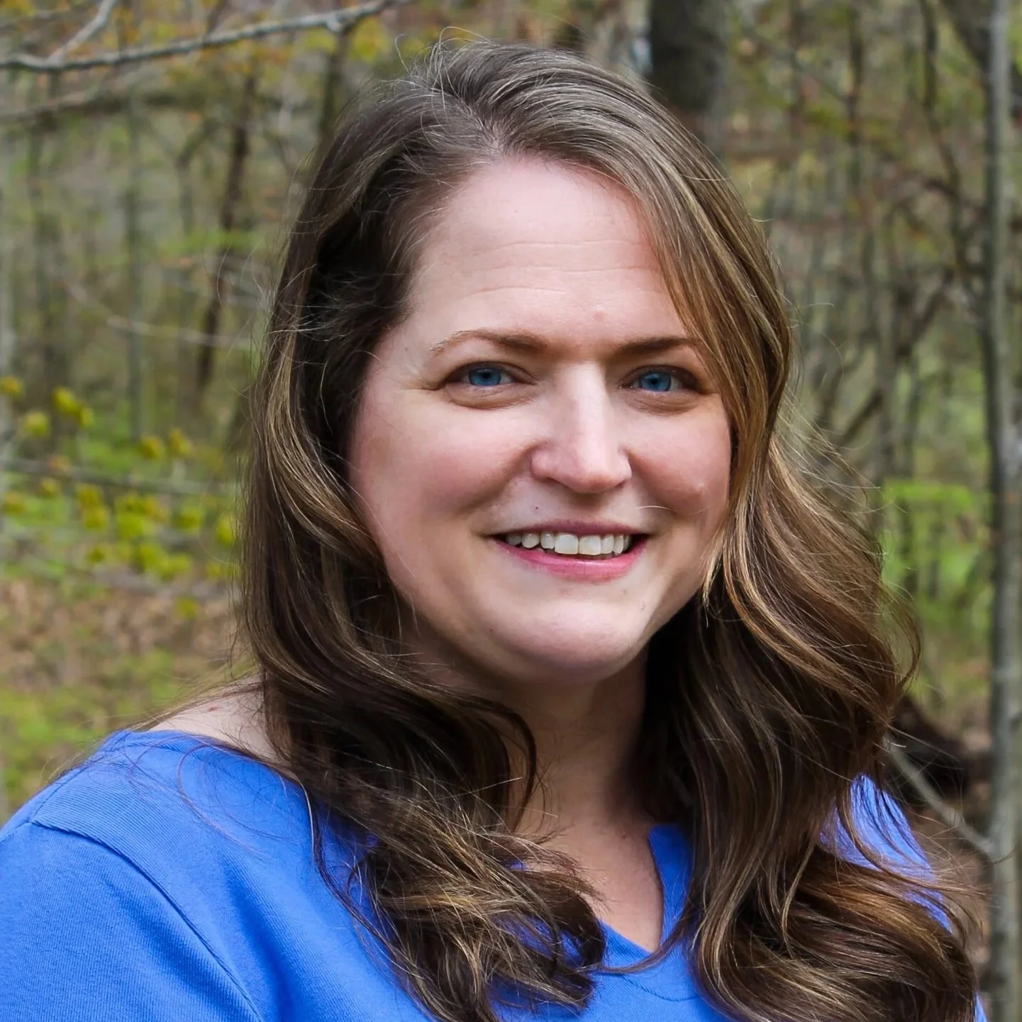 A woman with long brown hair, smiling, against a background of trees.