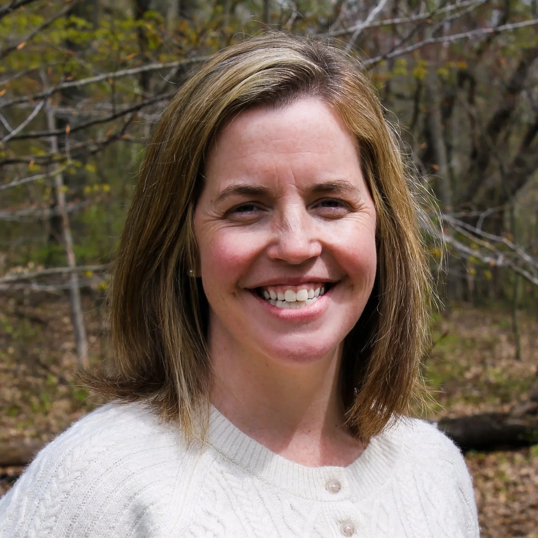 A smiling woman with shoulder length blonde hair, against a background of trees. 