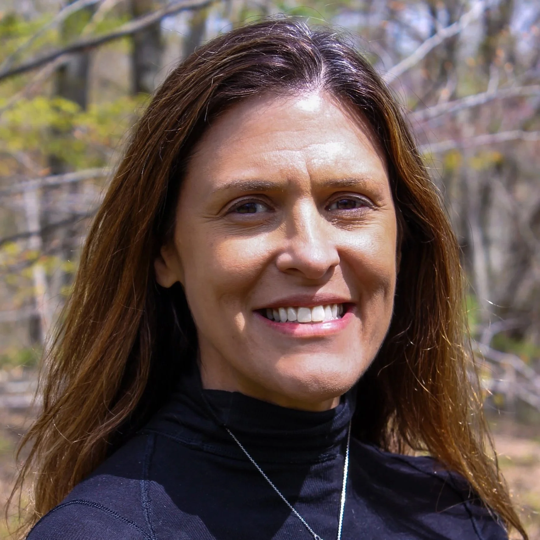 A woman with long brown hair and a black turtleneck smiling, standing outdoors.
