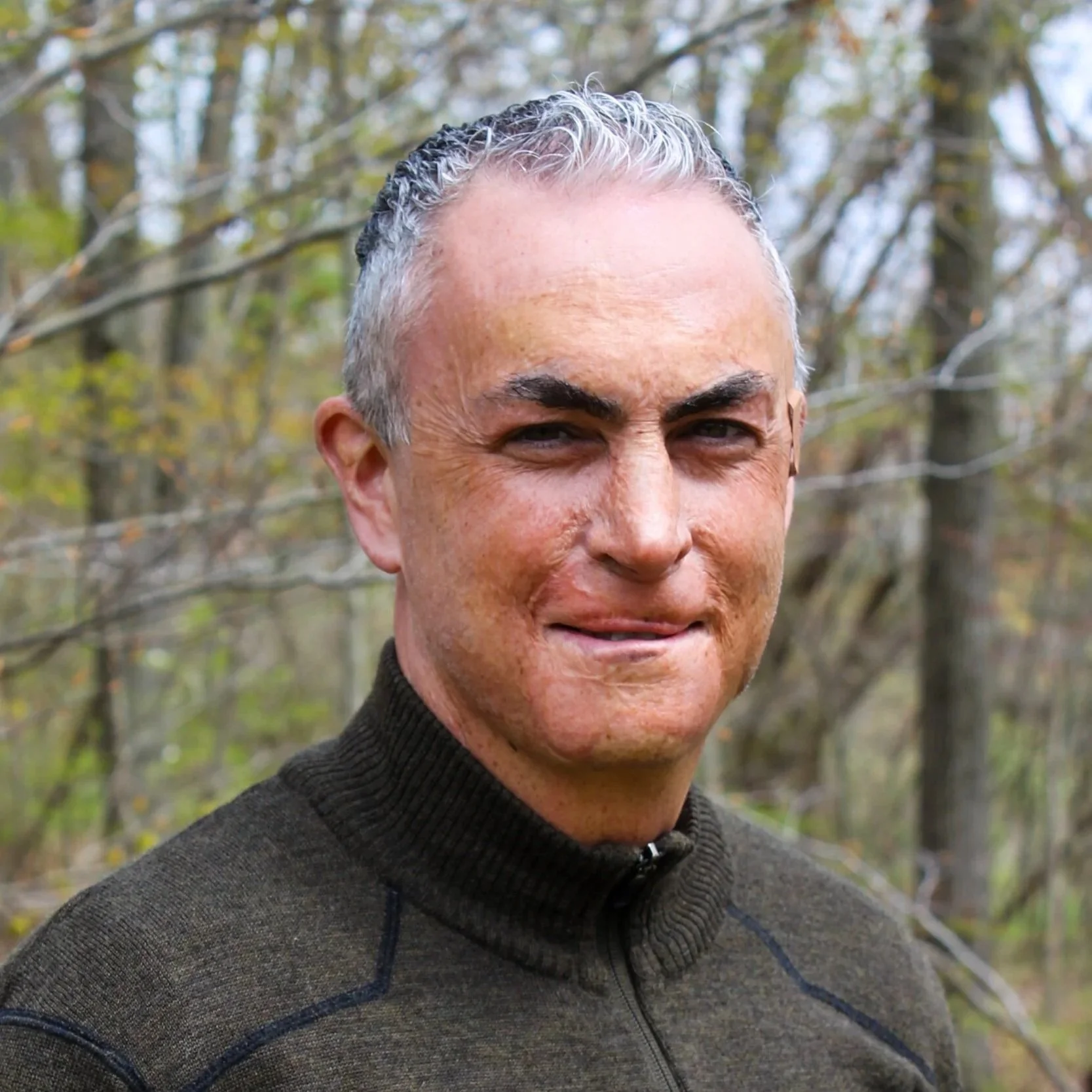 A man with short gray hair, smiling, against a background of trees. 