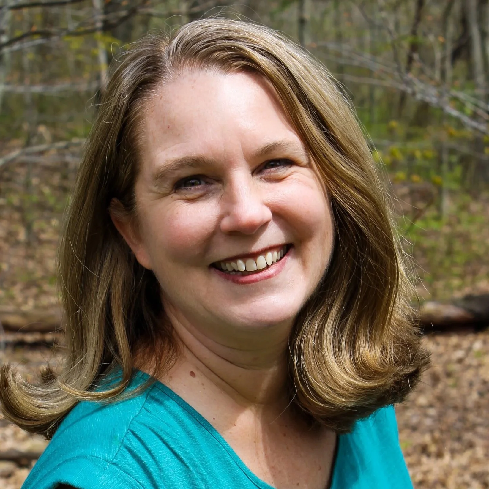 A woman with shoulder-length blonde hair, smiling, against a background of trees.