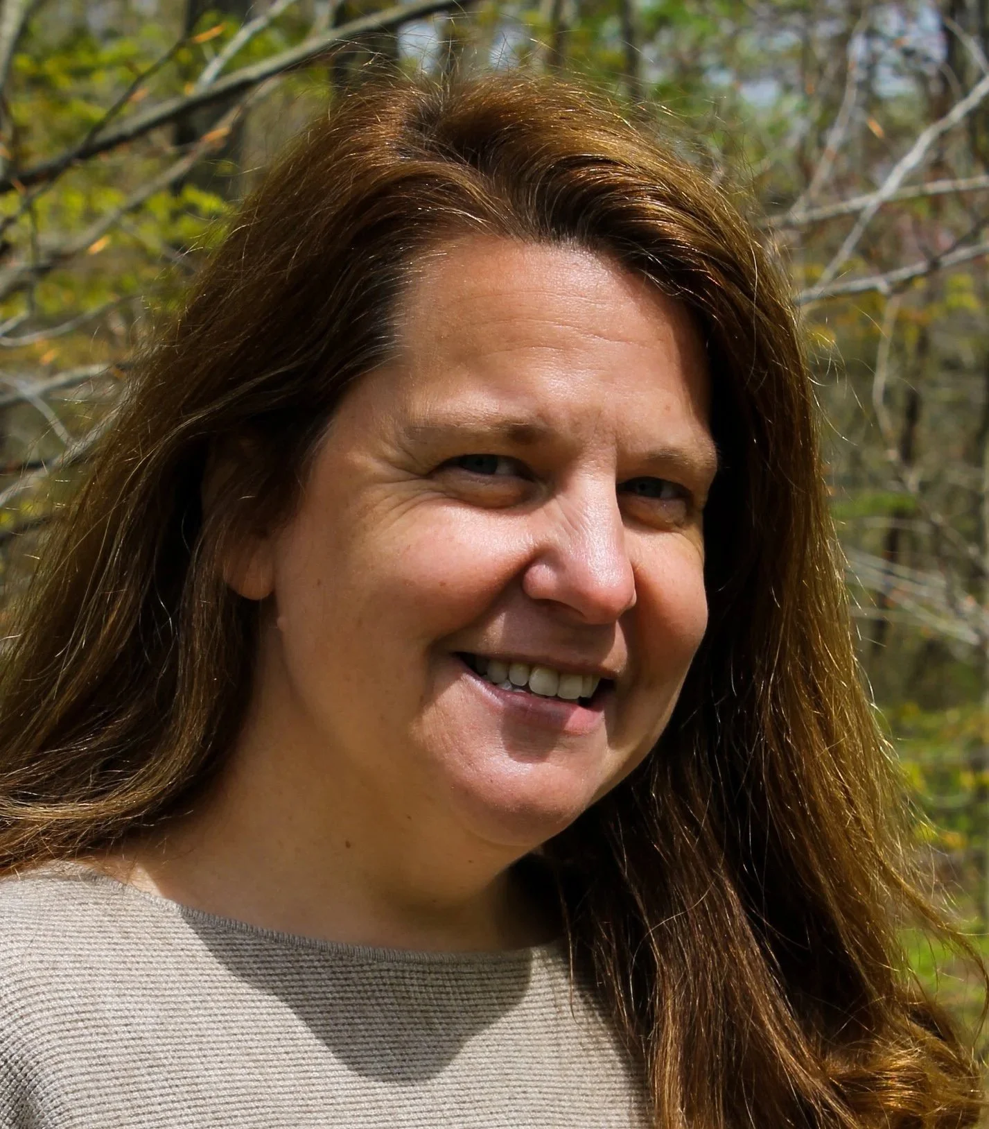 A woman with long brown hair, smiling, against a background of trees.