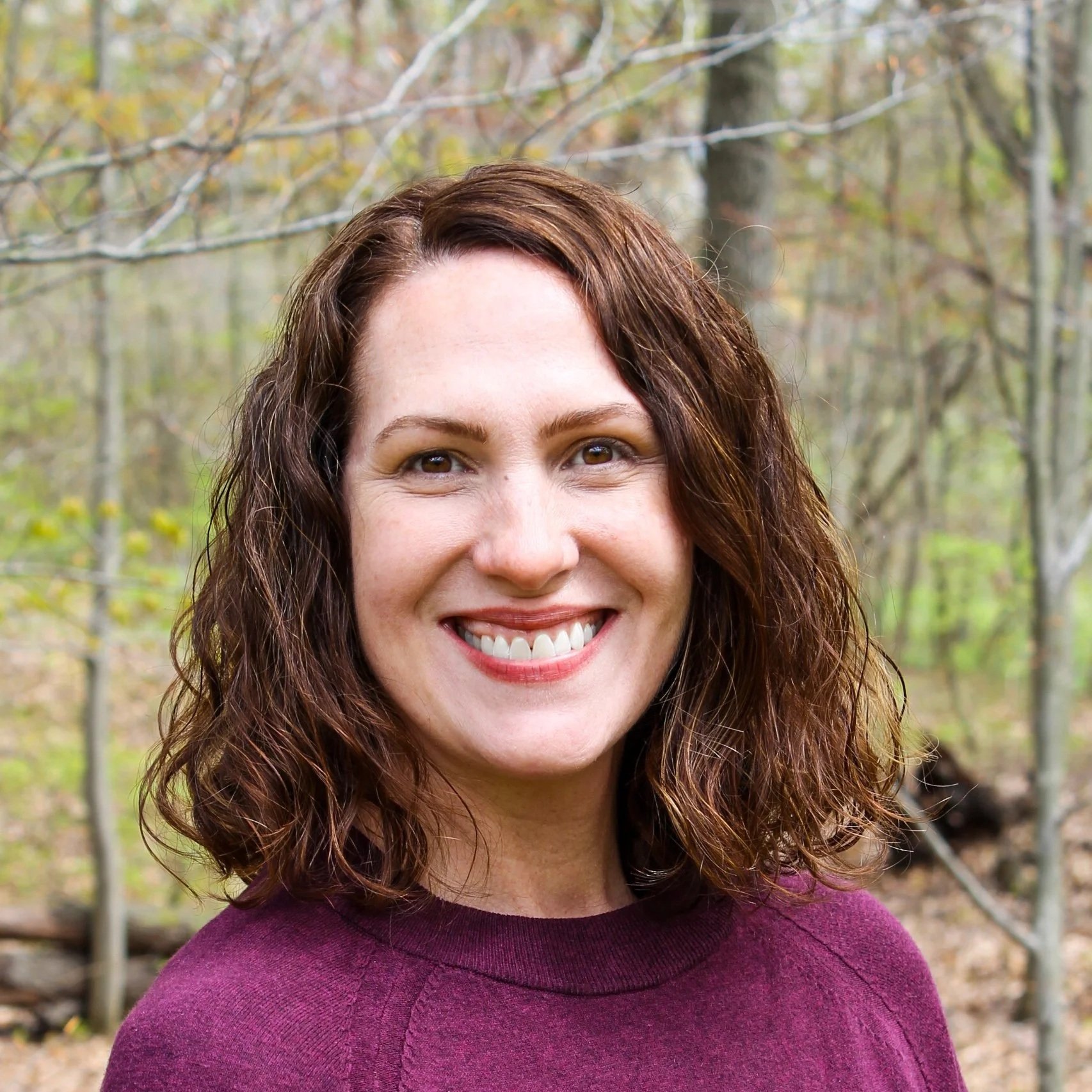 A woman with shoulder-length brown hair, smiling, against a background of trees. 