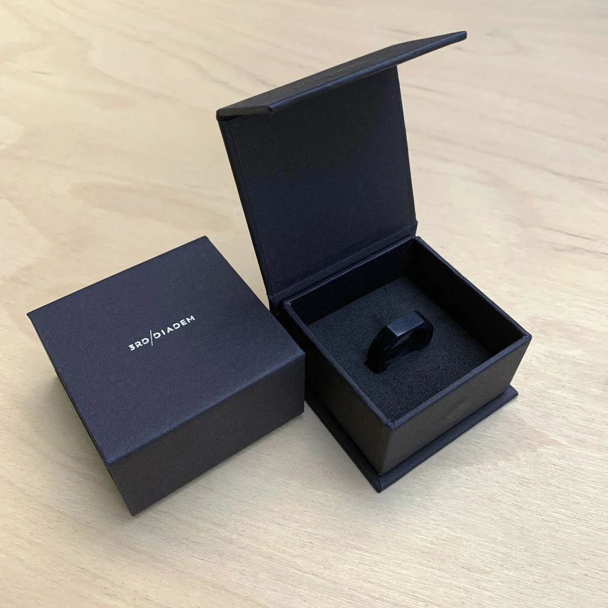 A minimalist design black ring inside a black jewelry box with a foam insert, next to a matching black outer box labeled "3RD/DIADEM" on a light wooden surface.
