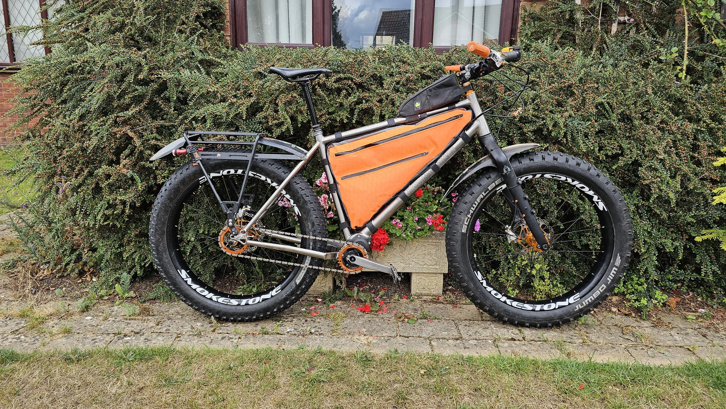 A mountain bike with fat tires parked in front of a bush with pink and red flowers, next to a brick house with windows.