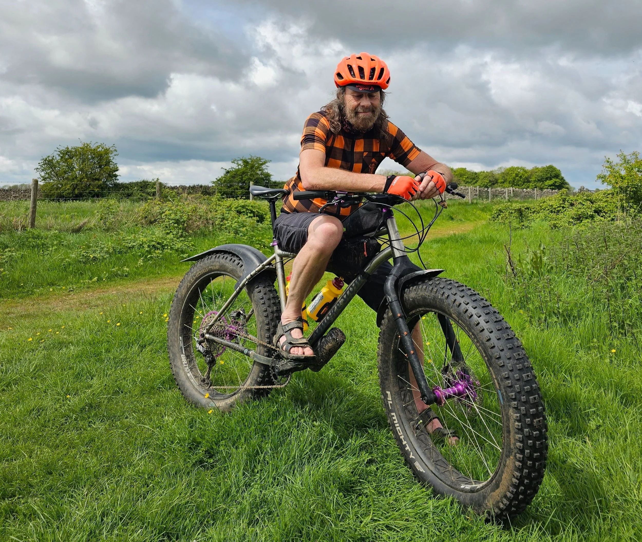 Graham Foot Bicycle Lover smiling with his Smokestone BowTi Fatbike bike on a grassy field, wearing a orange bike helmet, plaid shirt, shorts, and sandals, with cloudy skies overhead.